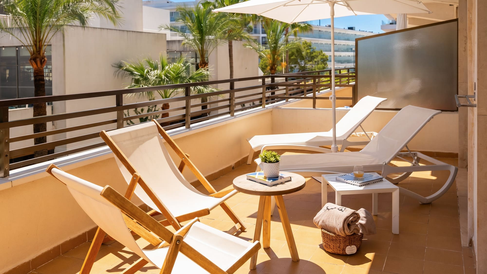 Sunny hotel balcony with white sun loungers, umbrella, and palm trees.