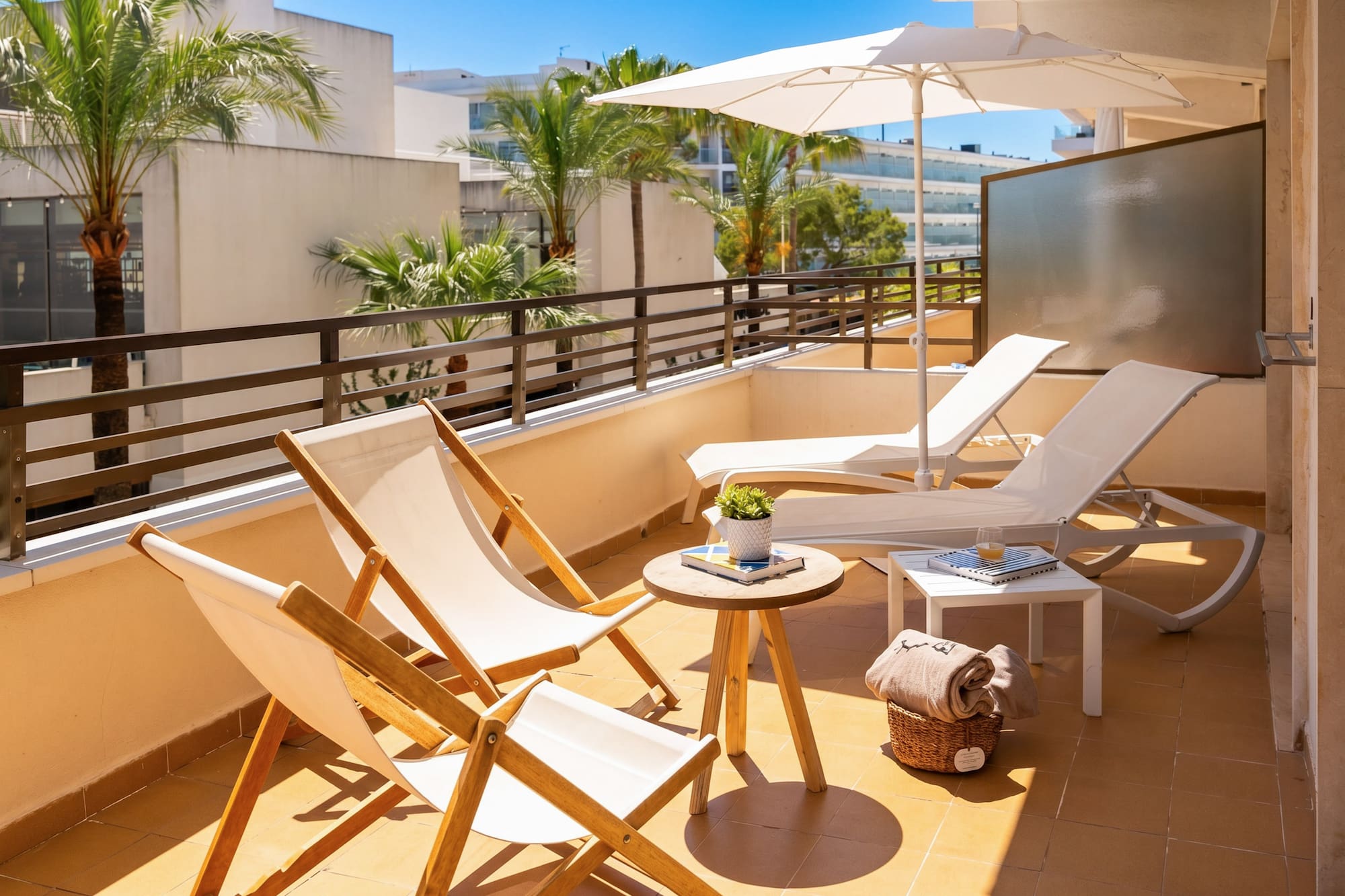 Sunny hotel balcony with white sun loungers, umbrella, and palm trees.