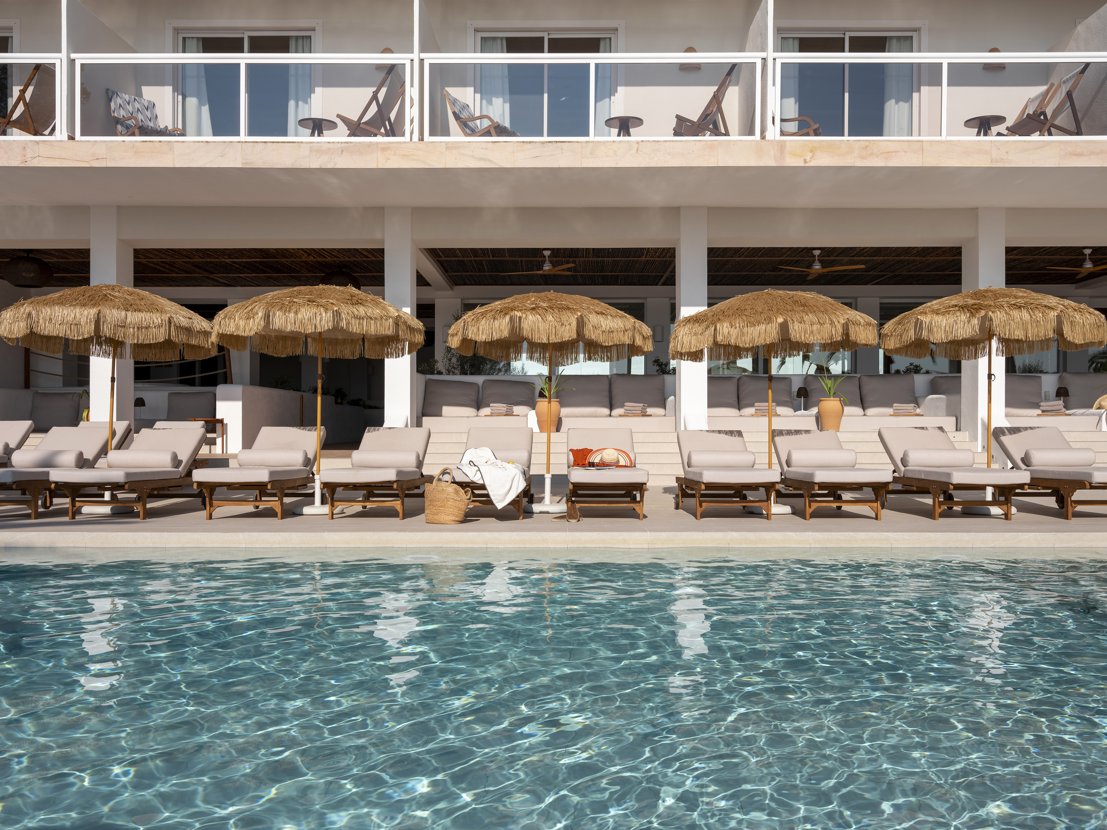 Resort swimming pool with straw umbrellas, lounge chairs, and hotel balconies.