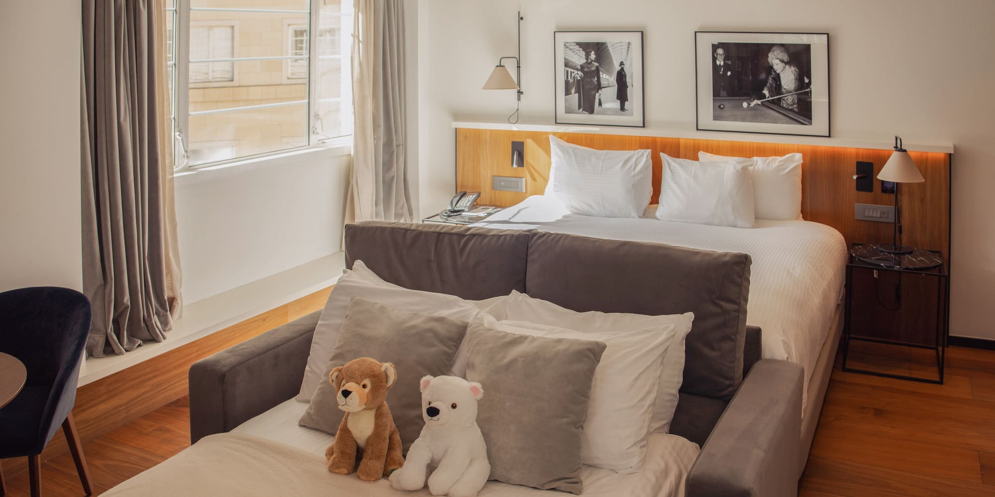 Modern hotel guest room with queen bed, sofa bed, and two plush teddy bears.