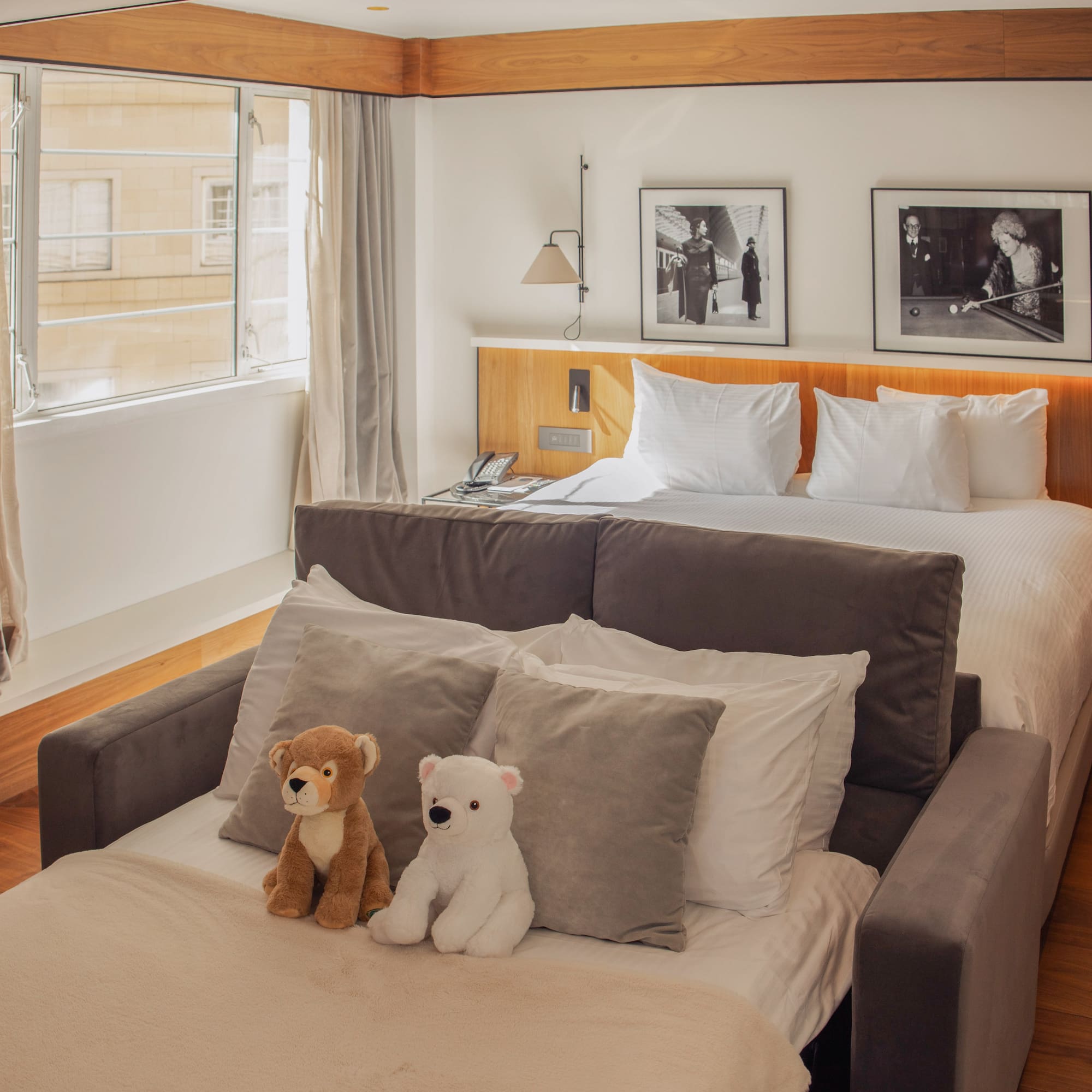 Modern hotel guest room with queen bed, sofa bed, and two plush teddy bears.