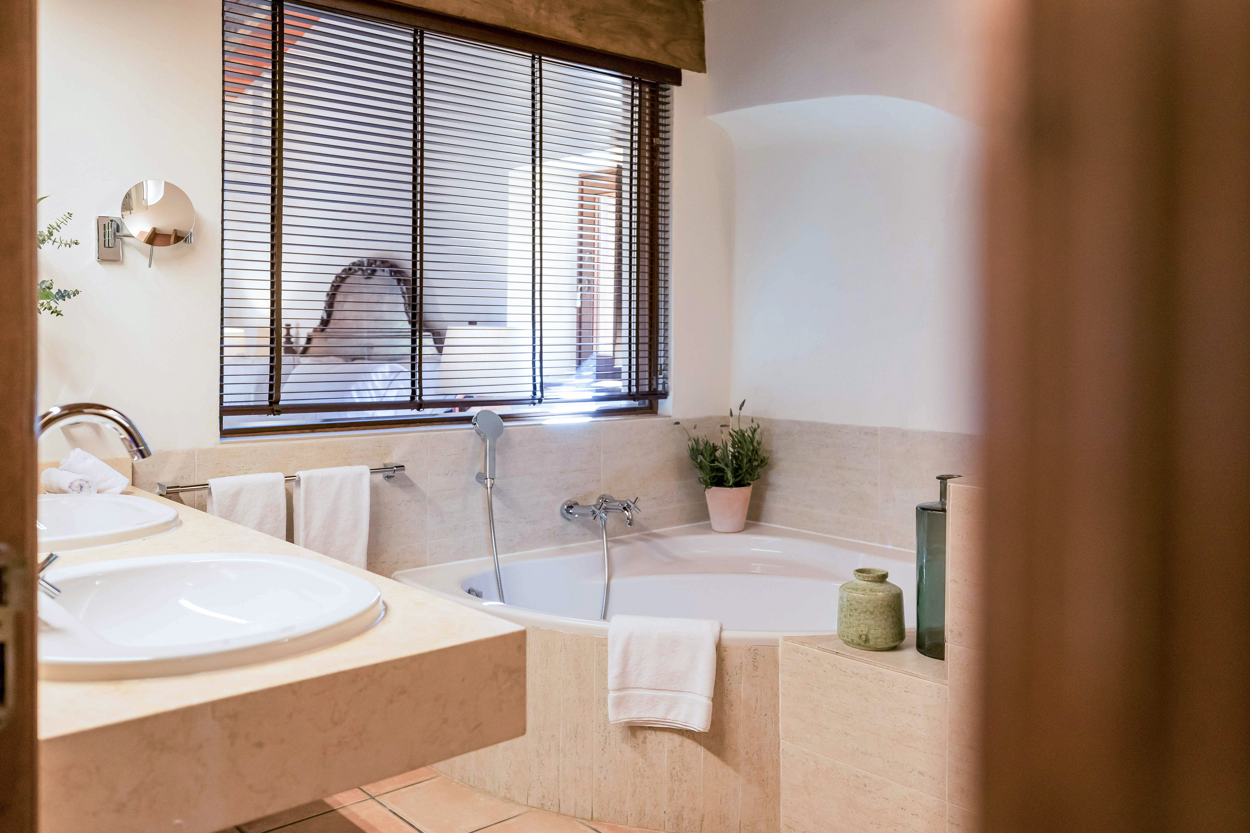 Bright hotel bathroom with double vanity, corner tub, wooden blinds.