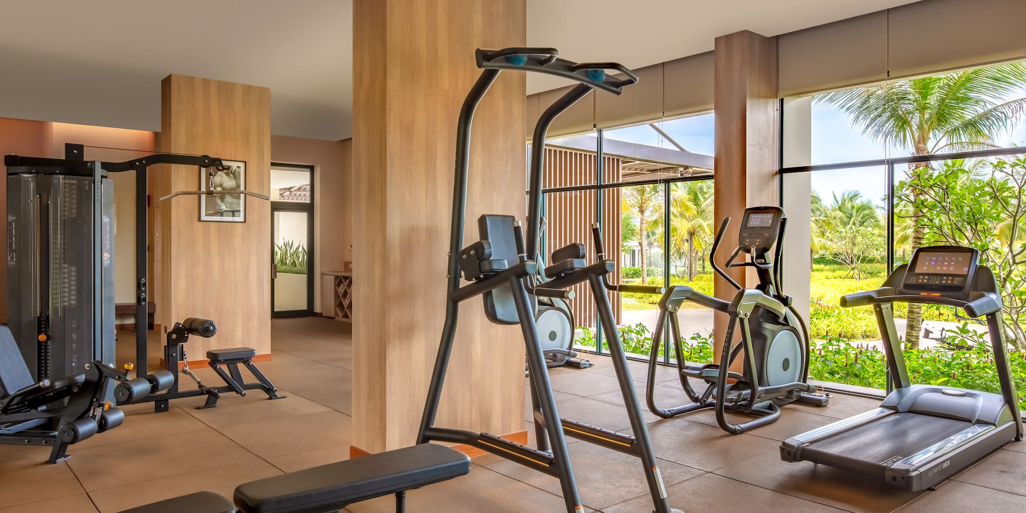 Modern gym with treadmill, elliptical, weight machines, and tropical outdoor view.