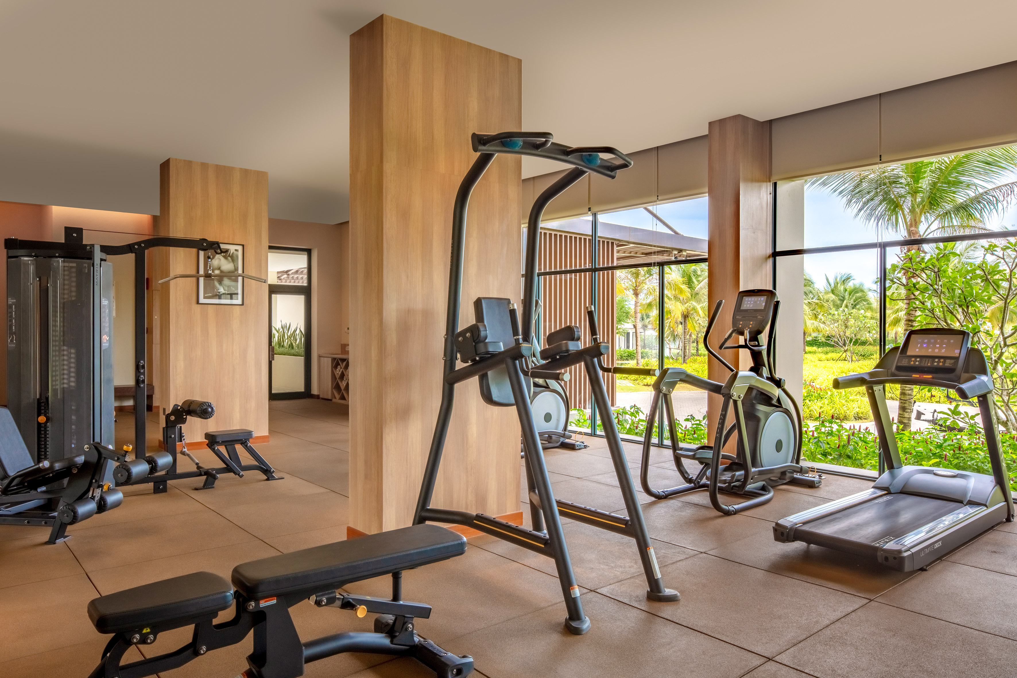 Modern gym with treadmill, elliptical, weight machines, and tropical view windows.