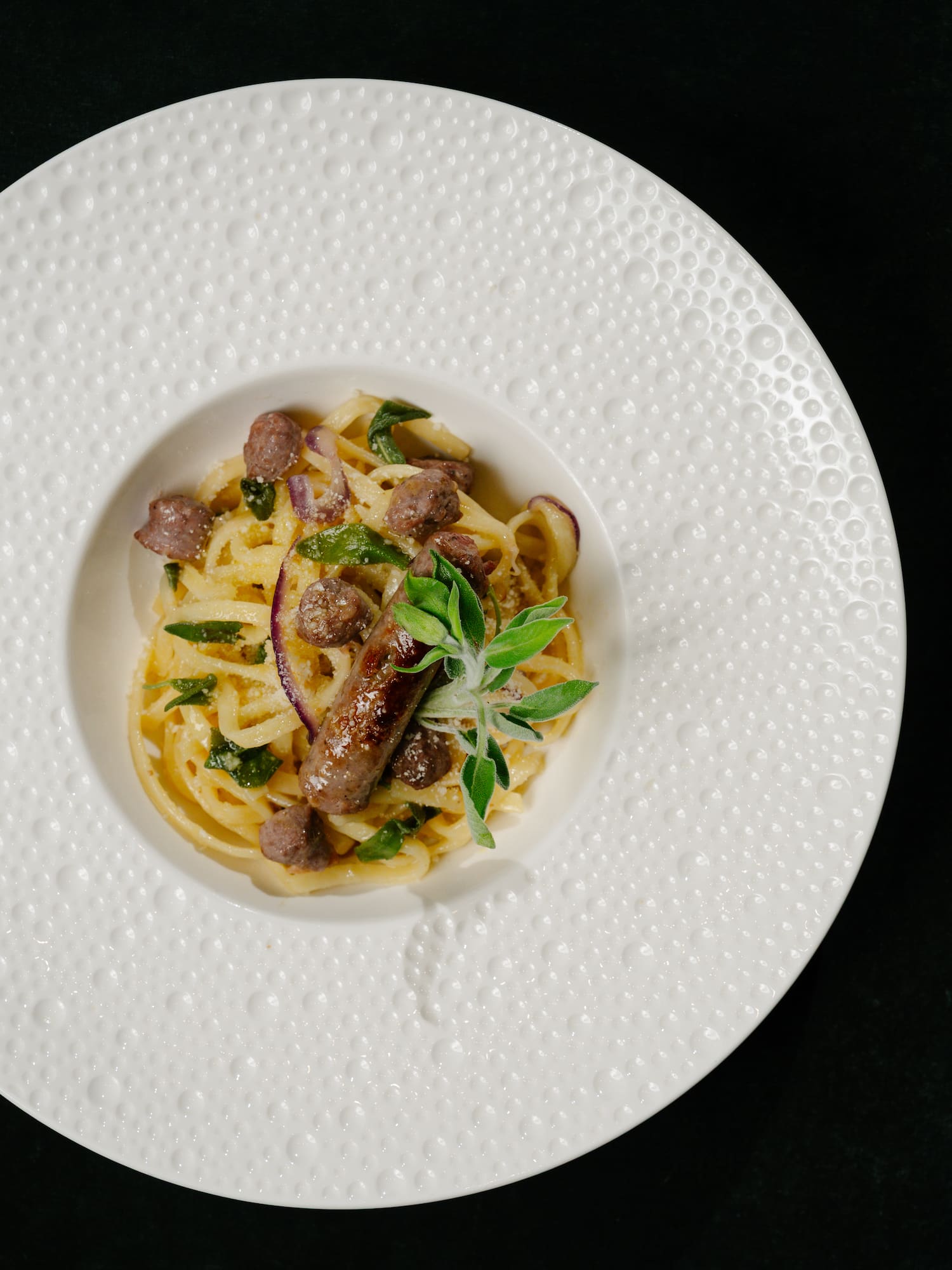 Sausage pasta with red onion, sage, and creamy sauce in a textured white bowl.