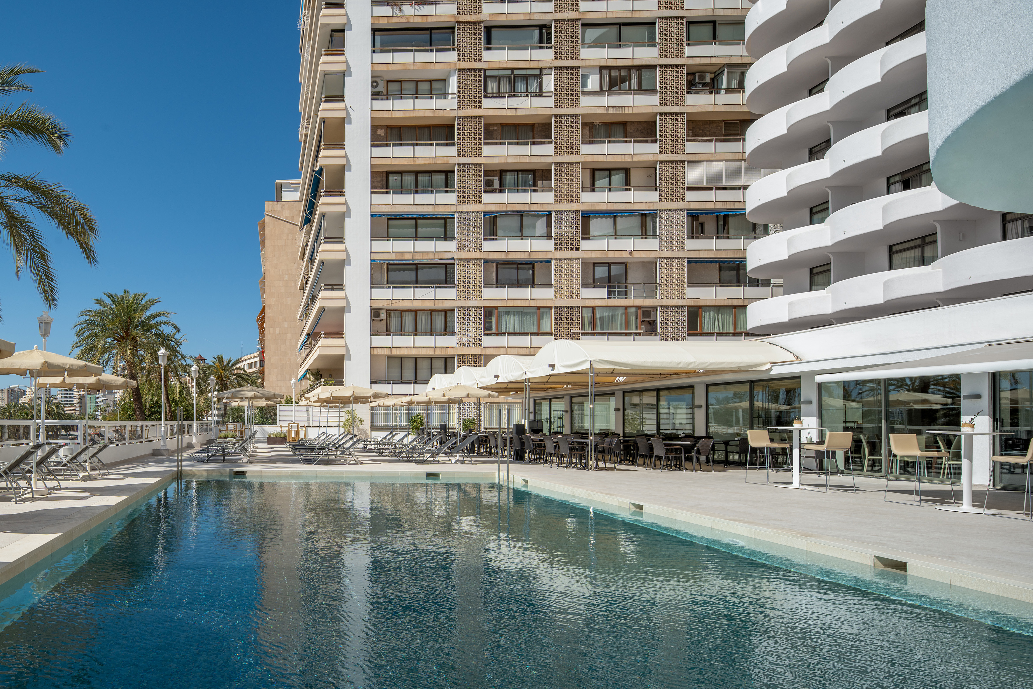 Sunny hotel pool area with modern buildings, outdoor dining, and palm trees.
