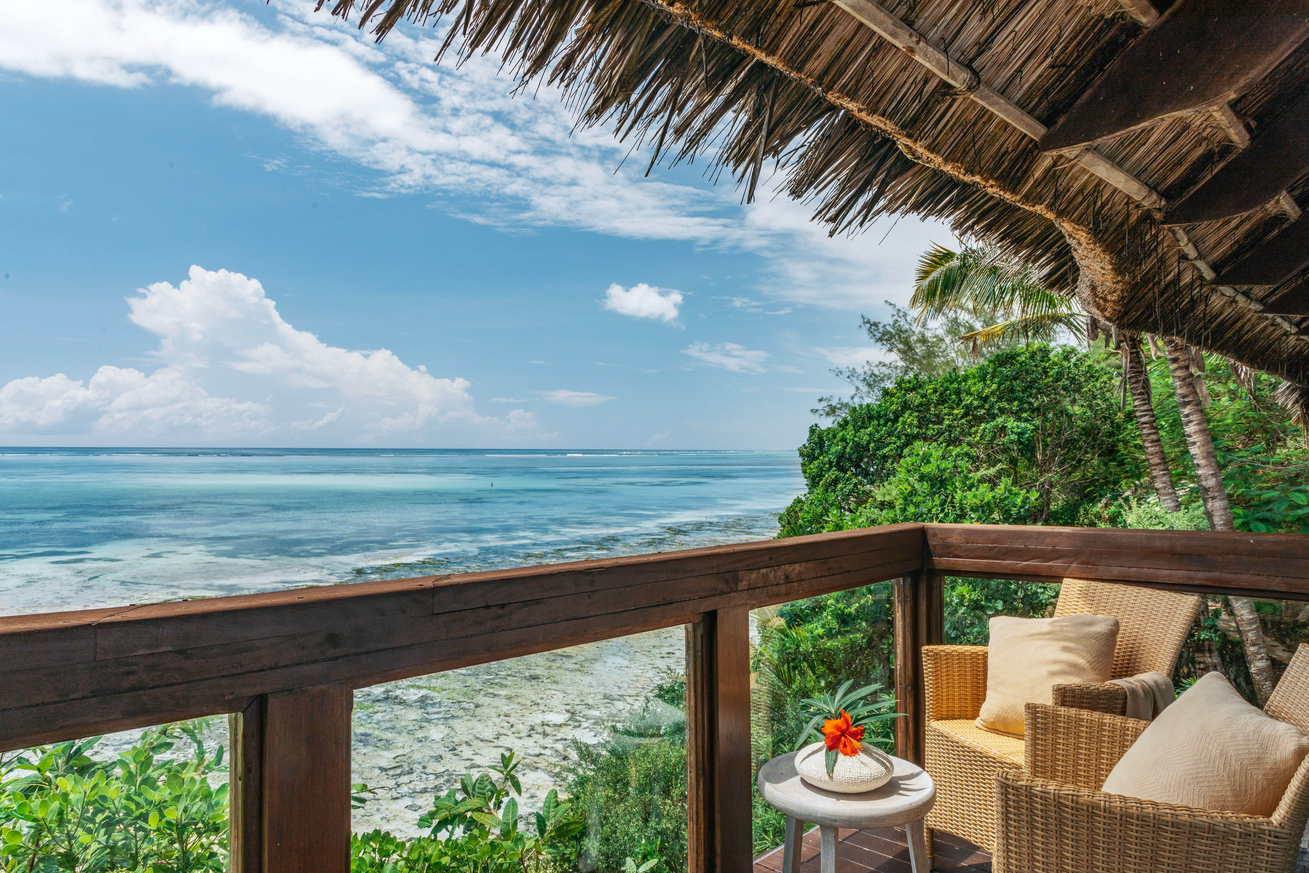 Tropical balcony with thatched roof overlooking turquoise ocean, lush foliage.