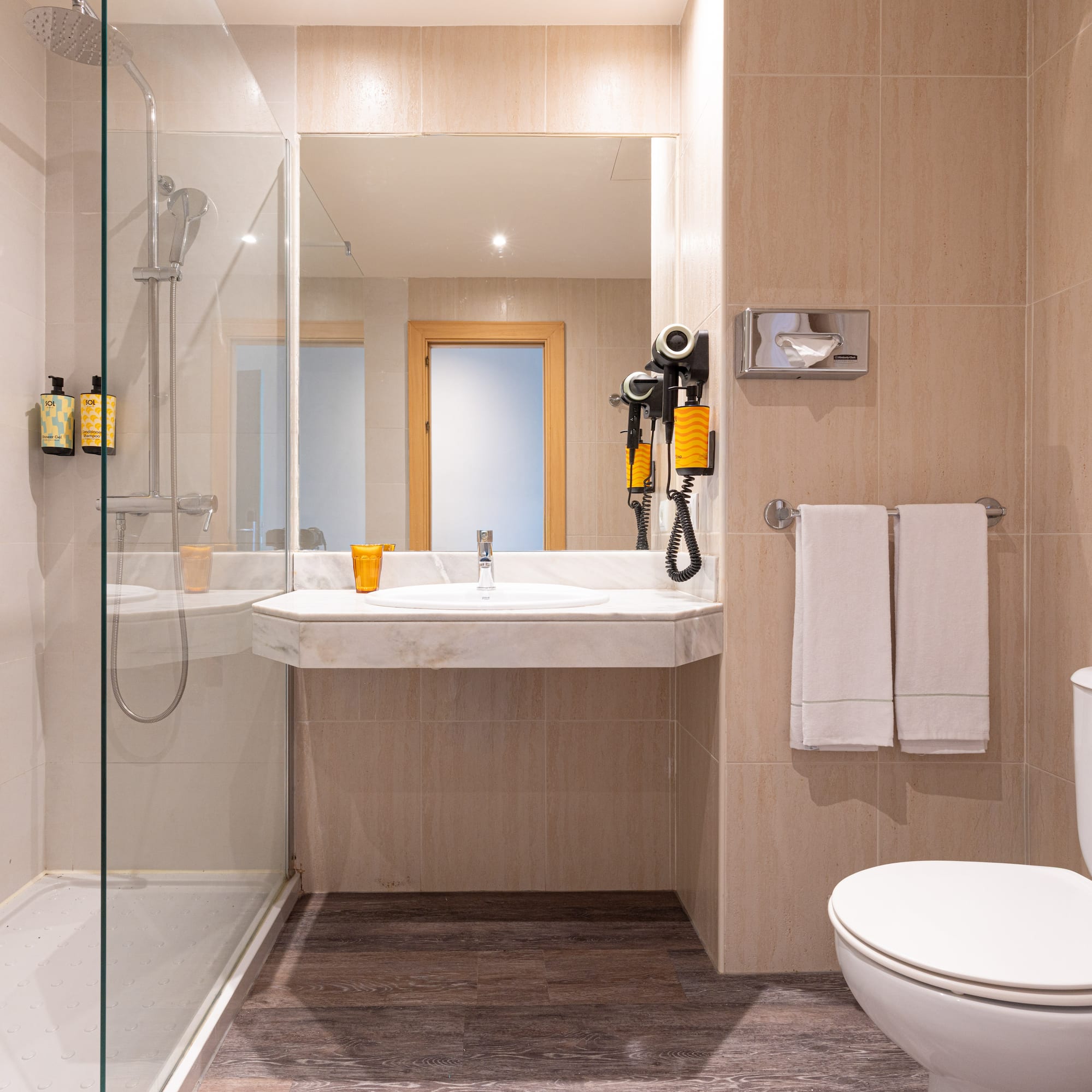 Baño de hotel con ducha de mampara, lavabo, espejo, inodoro y azulejos beige.