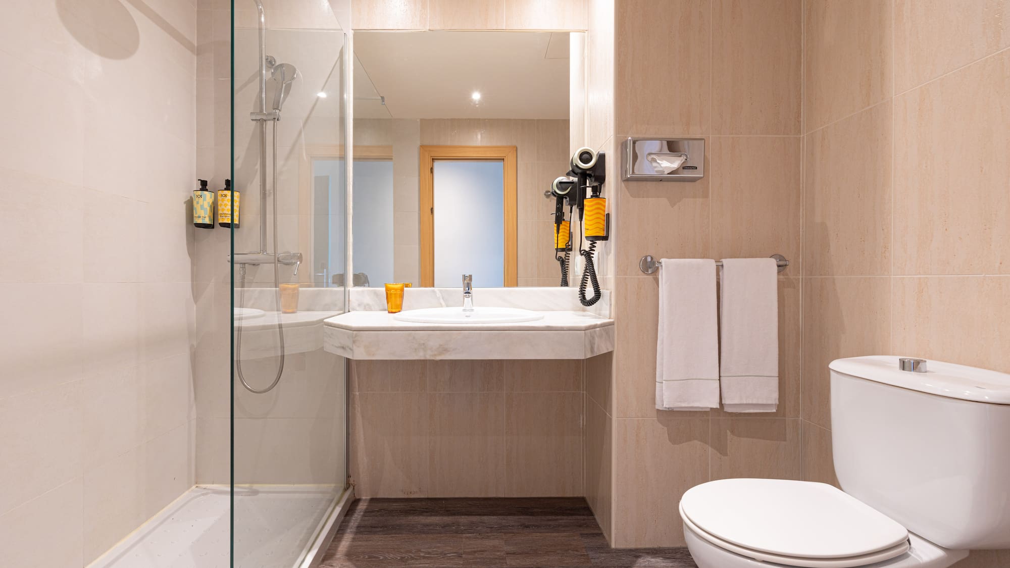 Baño de hotel con ducha de mampara, lavabo, espejo, inodoro y azulejos beige.