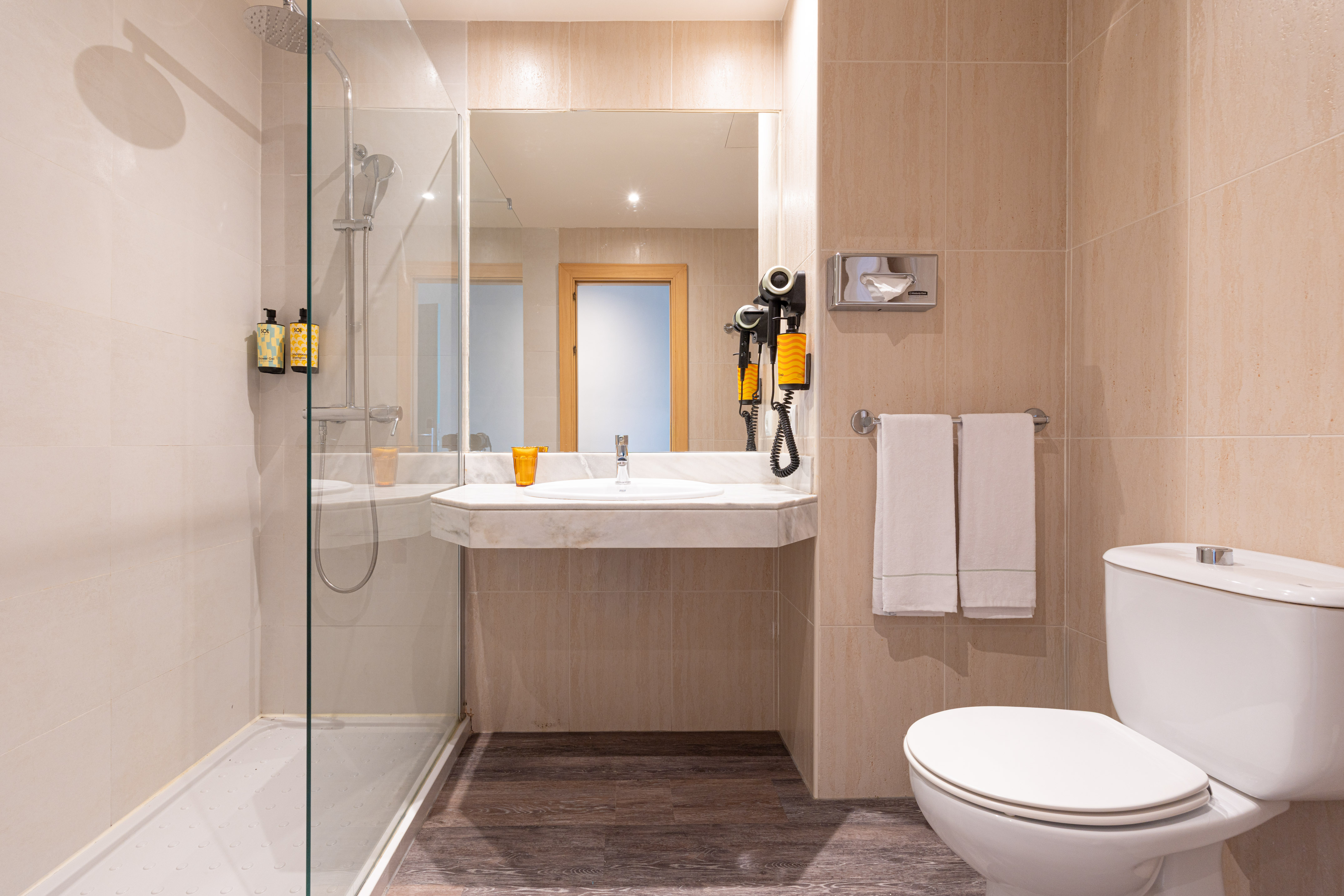 Baño de hotel con ducha de mampara, lavabo, espejo, inodoro y azulejos beige.