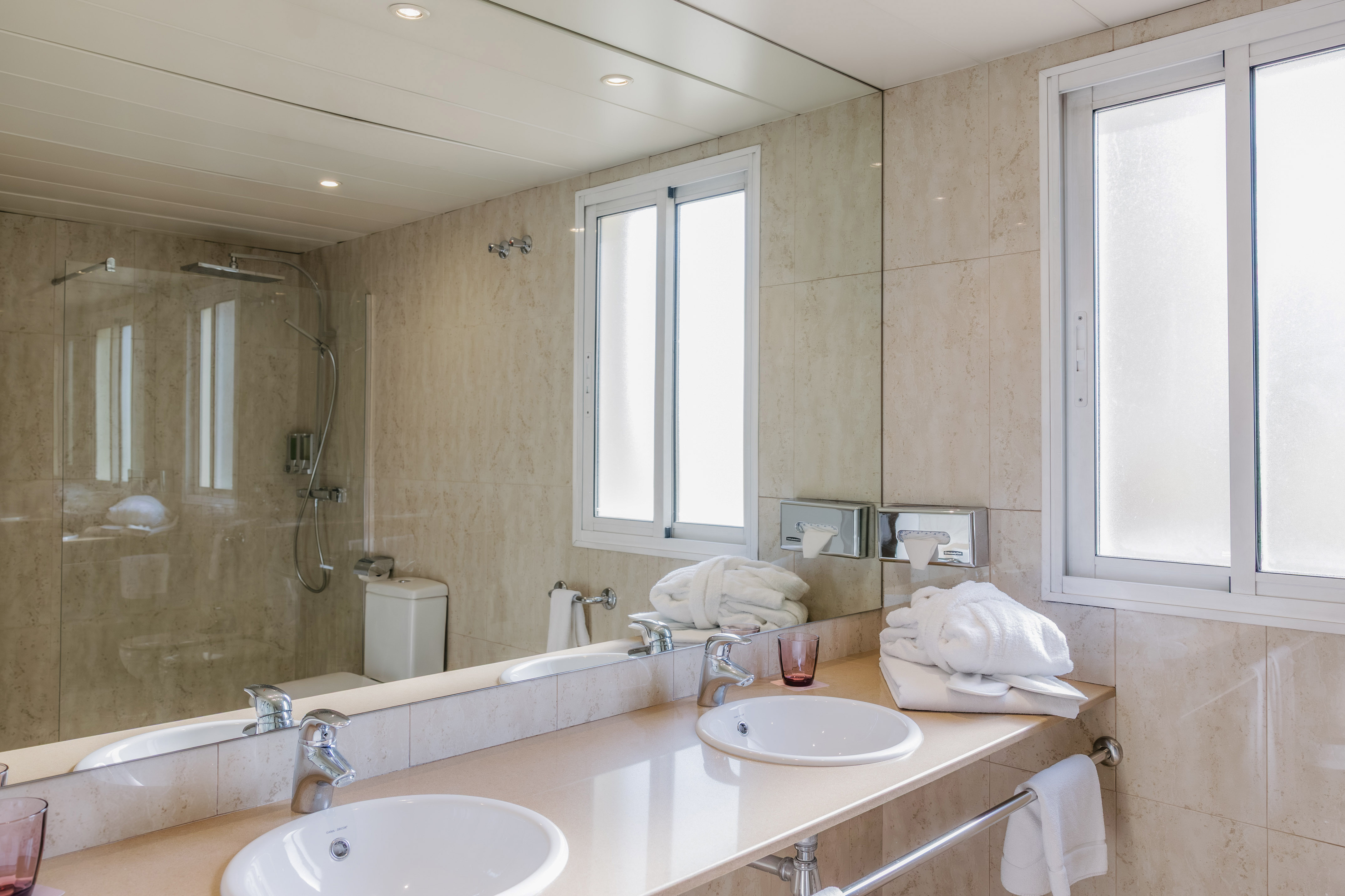 Modern hotel bathroom with double sinks, mirror, shower, and window.