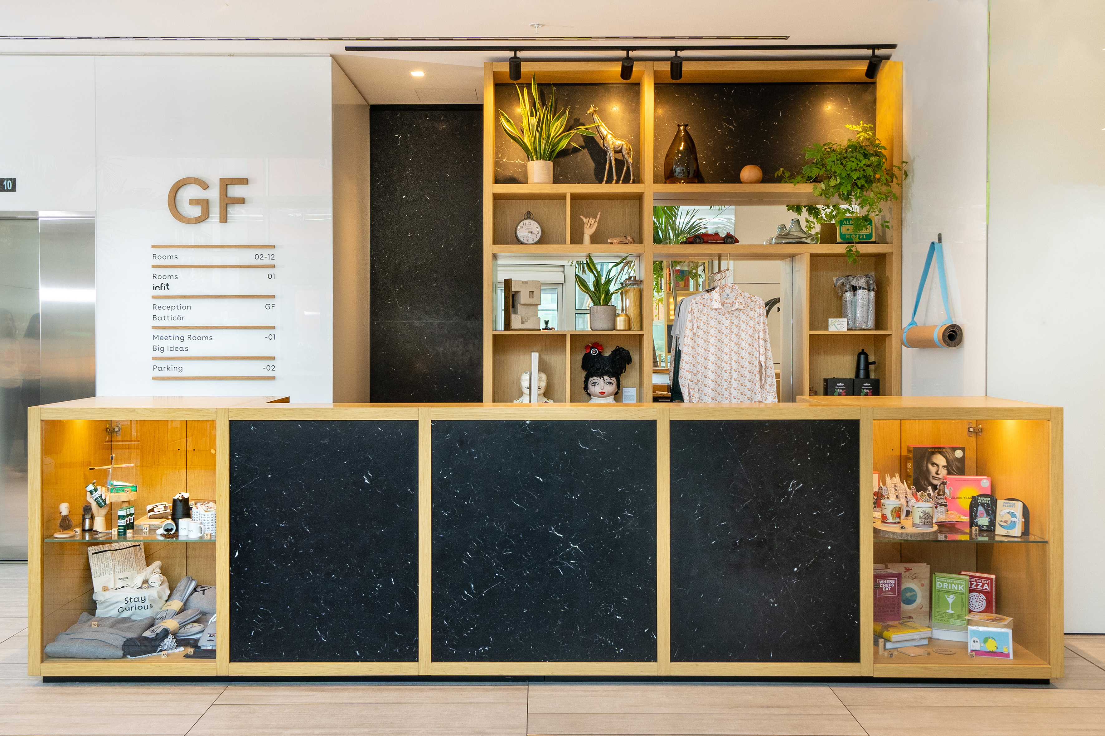 Modern hotel reception desk with wood, black marble, plants, and retail displays.