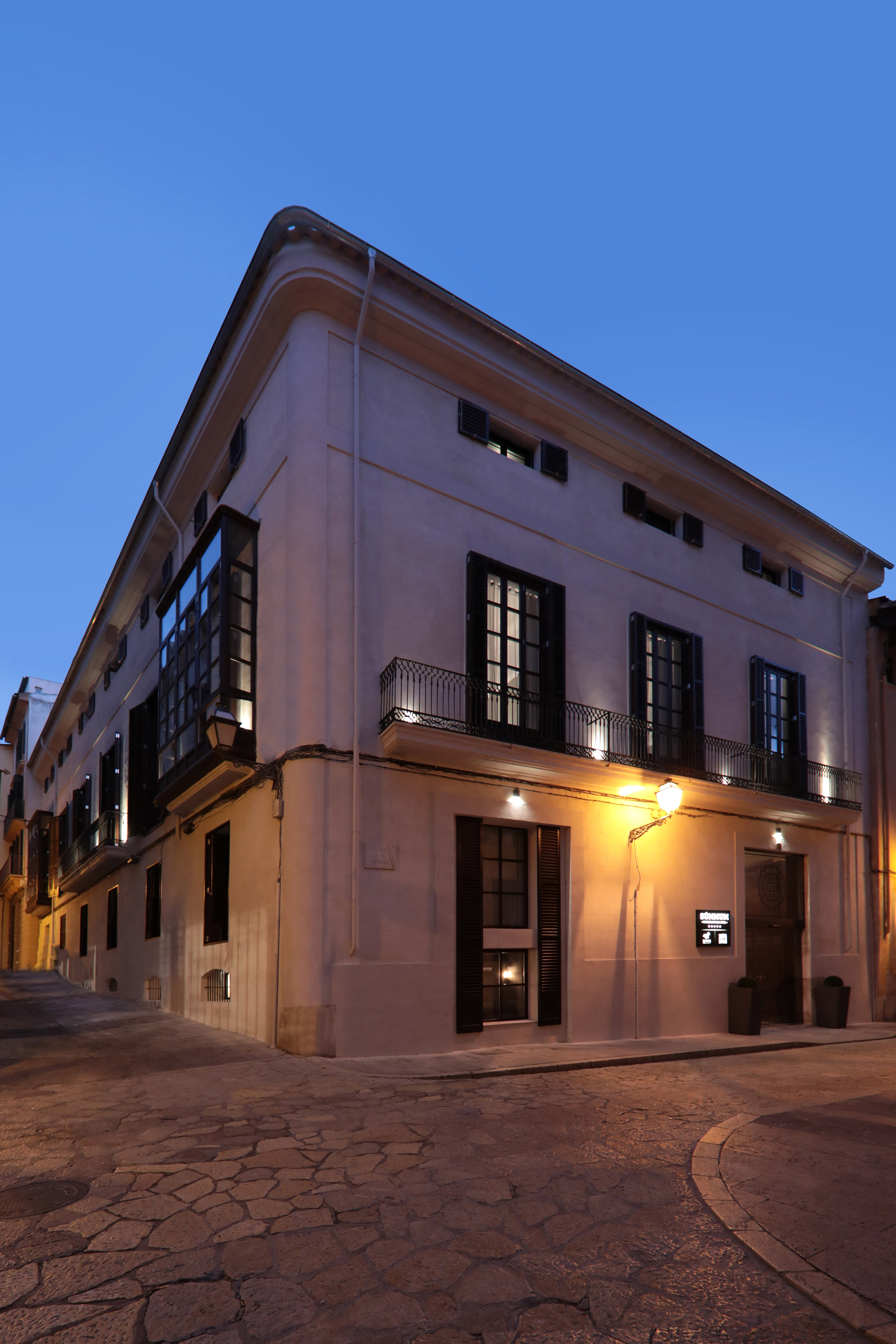 Illuminated corner building, dark windows, balconies, cobblestone street, at dusk.