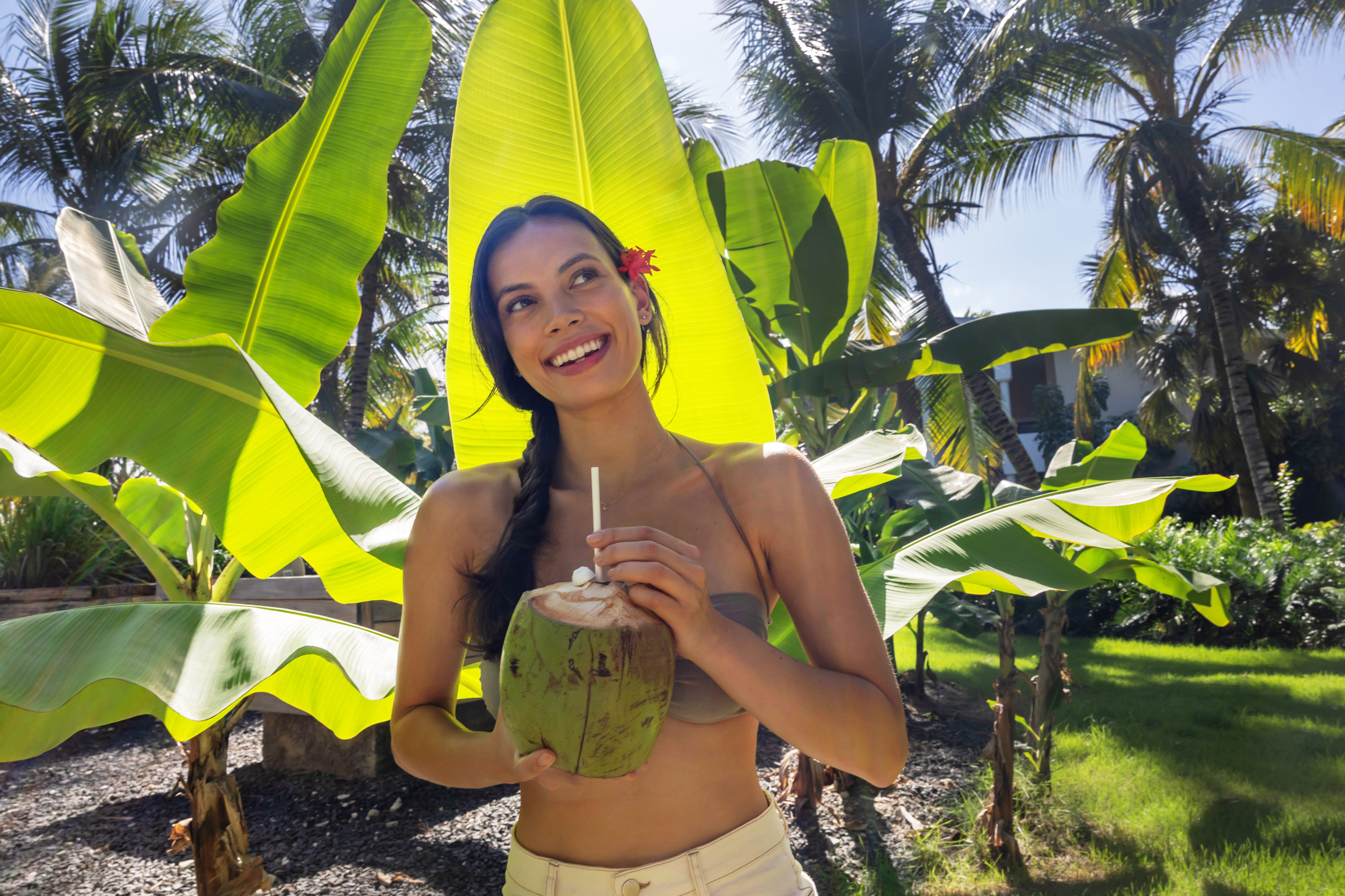 Smiling woman with coconut drink under bright banana leaves and palm trees.