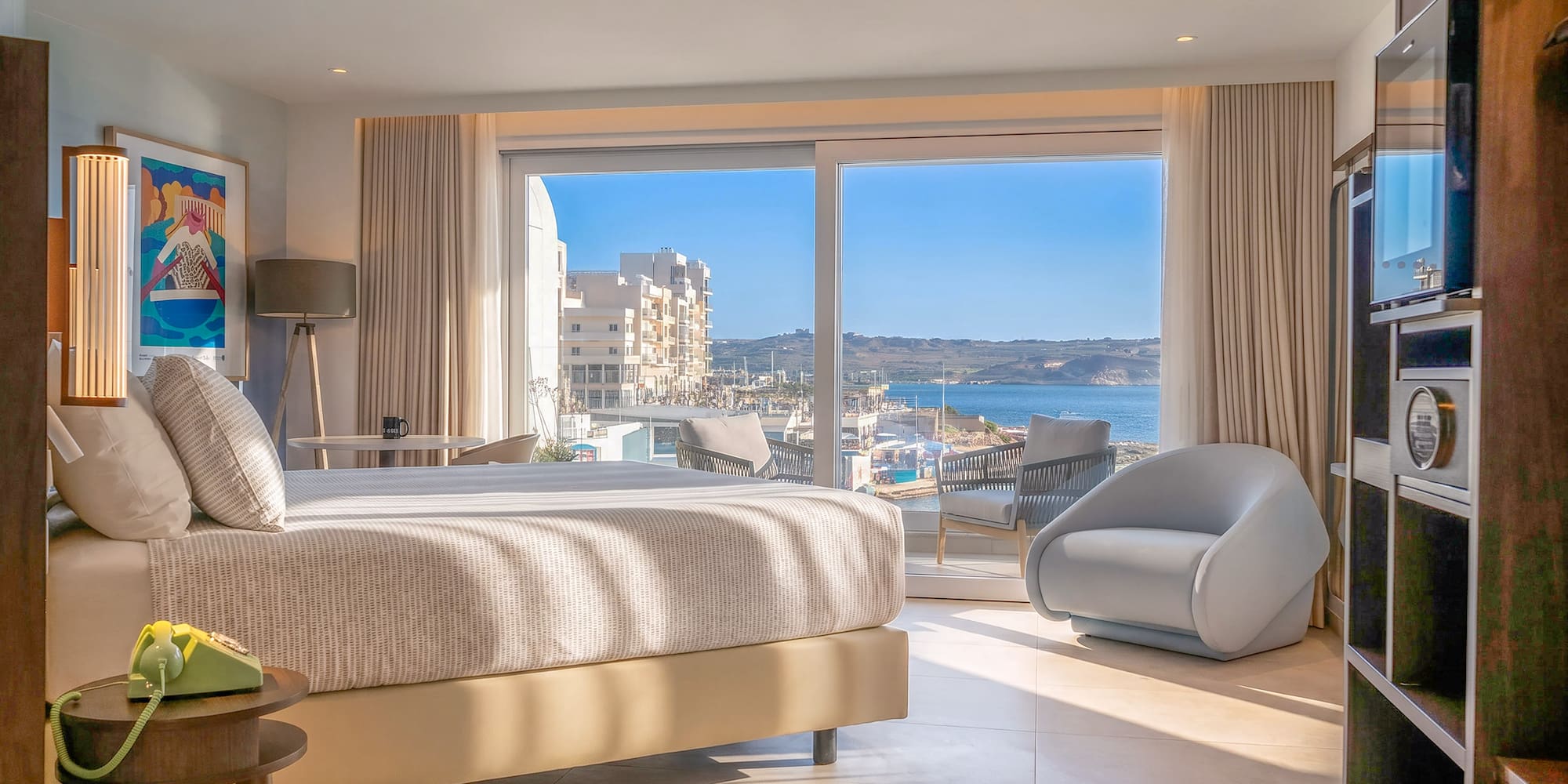 Modern hotel bedroom with large sea view window overlooking a sunny bay.