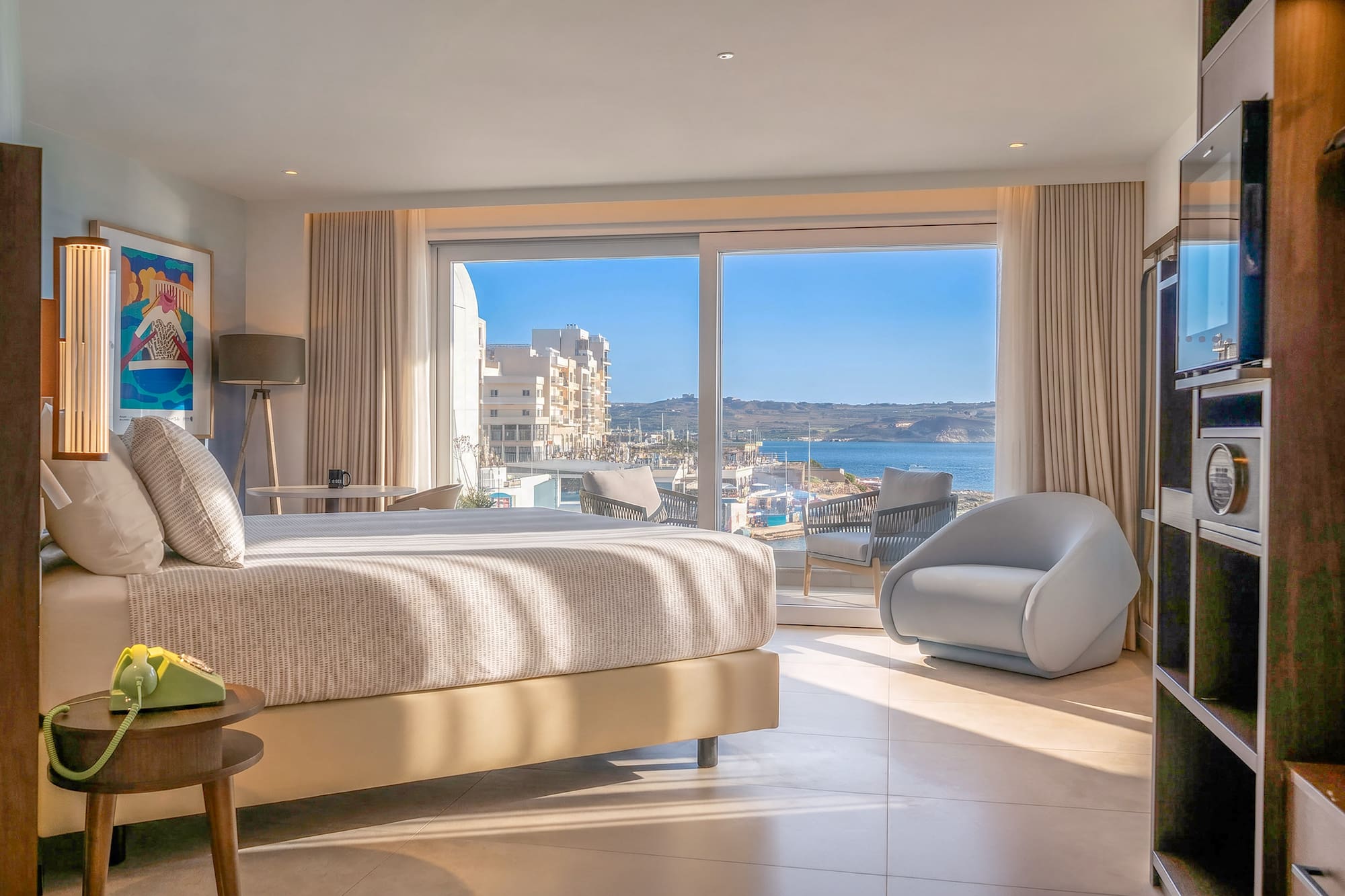 Modern hotel bedroom with large sea view window overlooking a sunny bay.