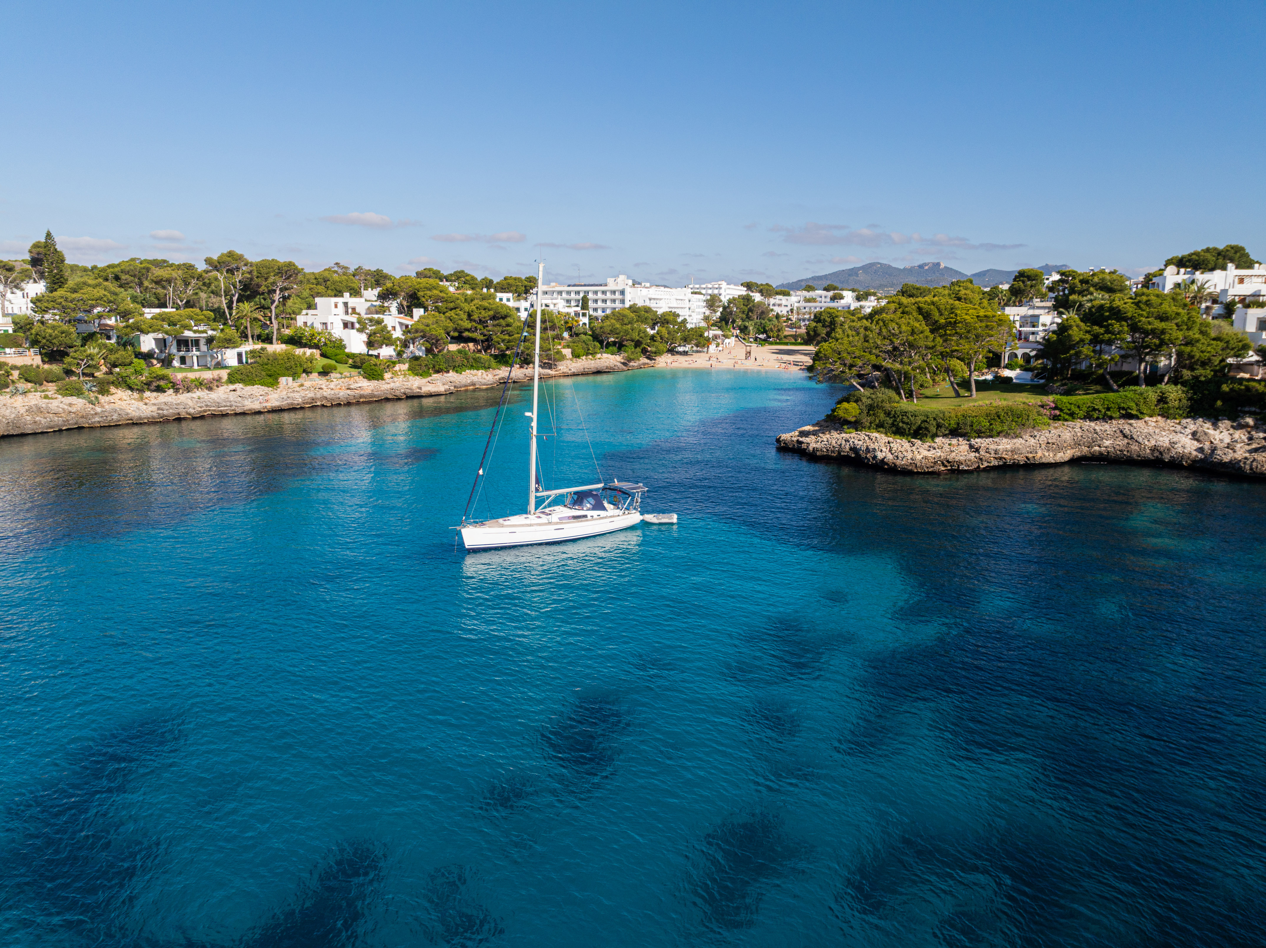 White sailboat in clear blue bay, sandy beach, coastal resort, pine trees.