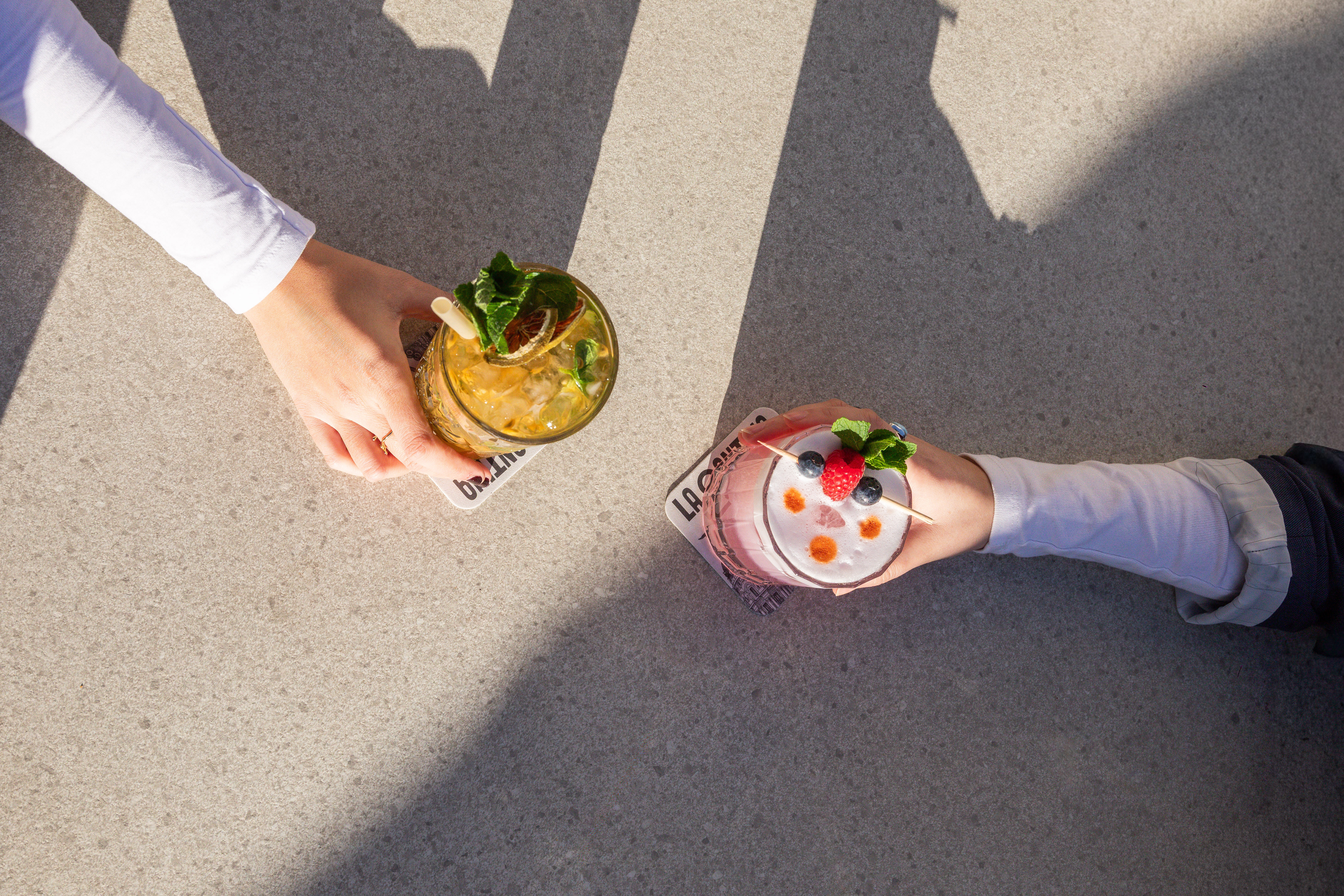 Hands holding two colorful fruity cocktails on a bright outdoor table.