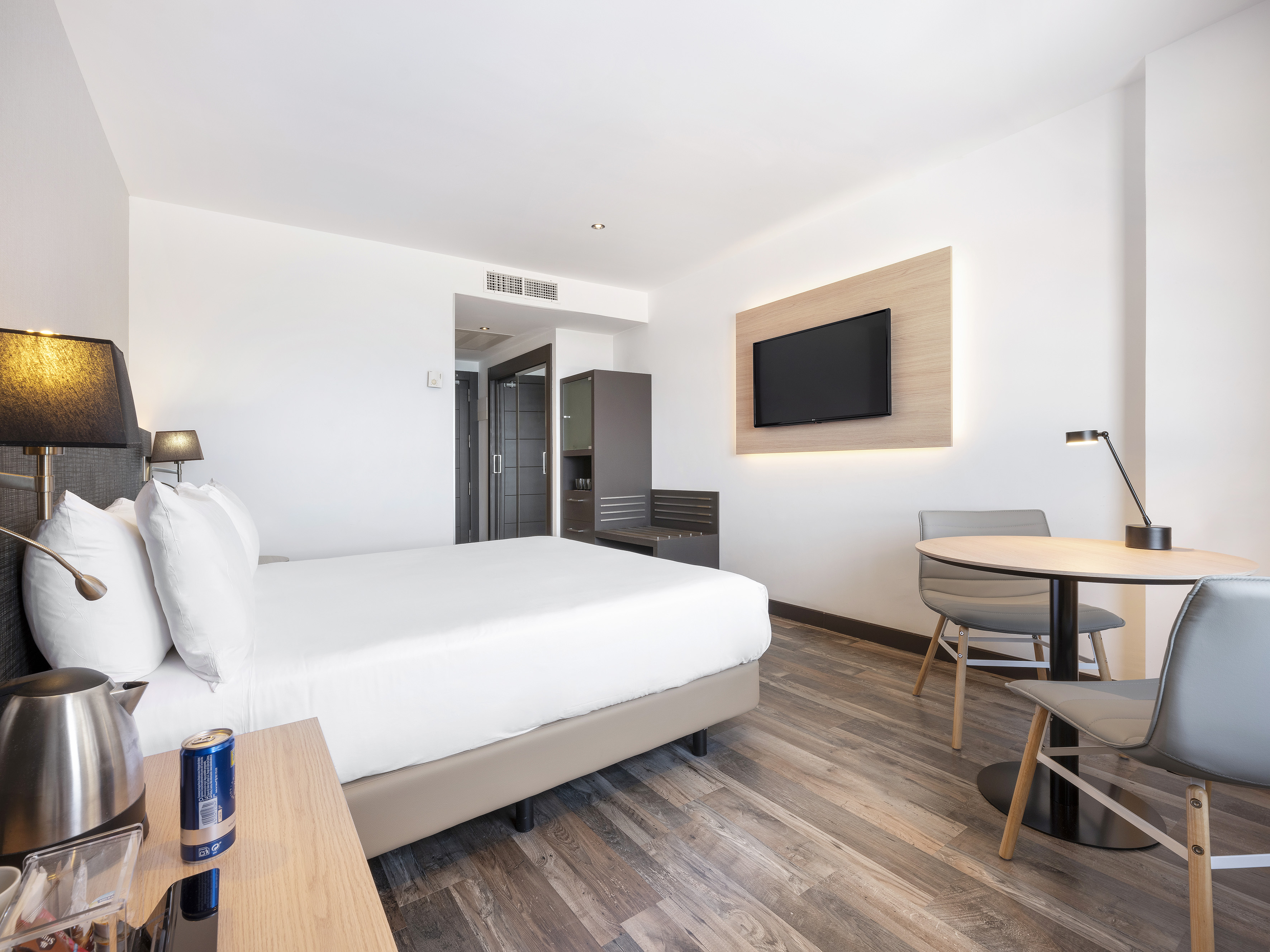 Modern hotel room with queen bed, wood floor, wall TV, and dining area.