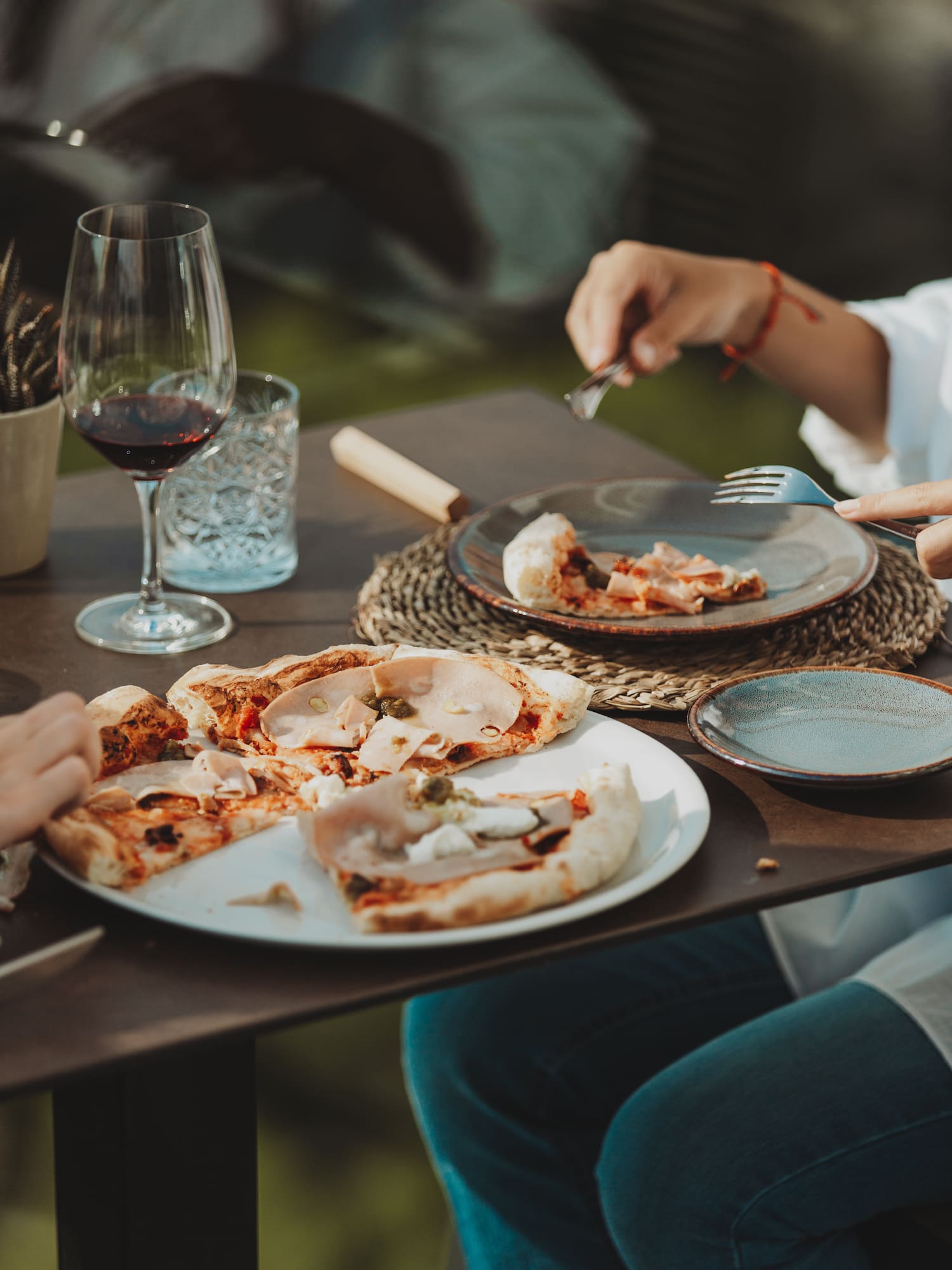 People eating pizza with ham, cheese, and red wine outdoors.