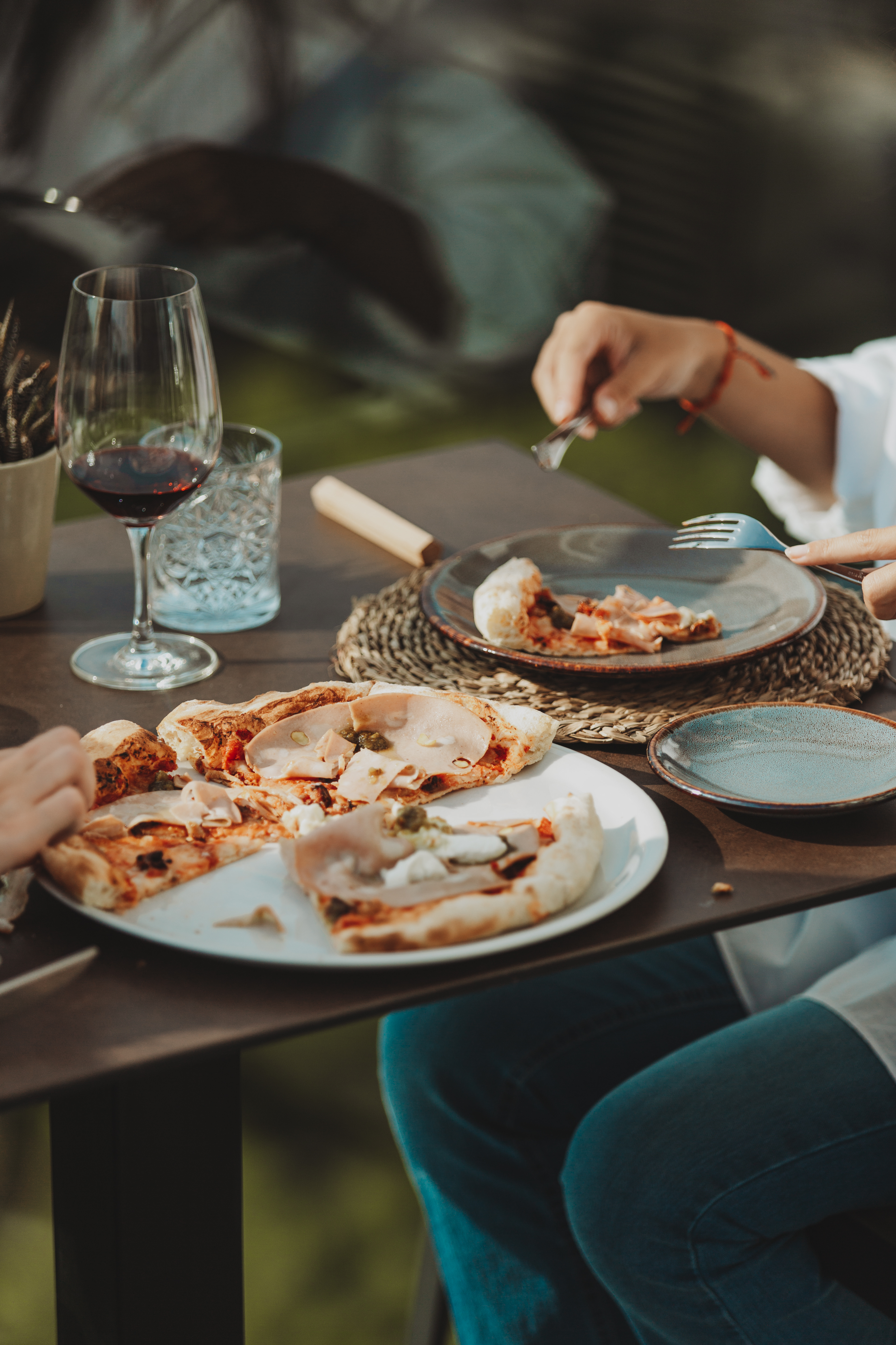 People eating pizza with ham, cheese, and red wine outdoors.