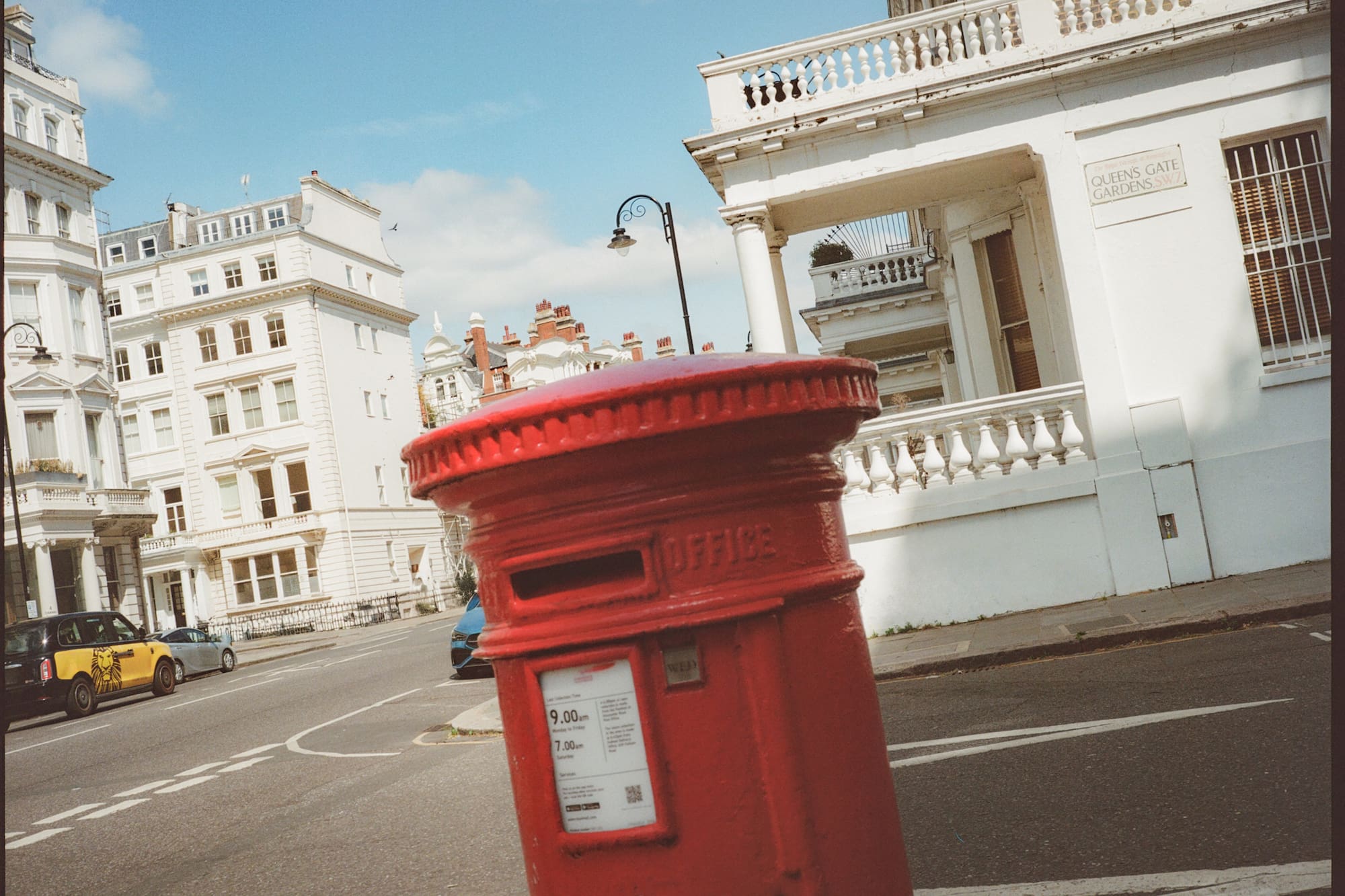 Buzón rojo de Londres en calle con edificios blancos, taxi y cielo azul.