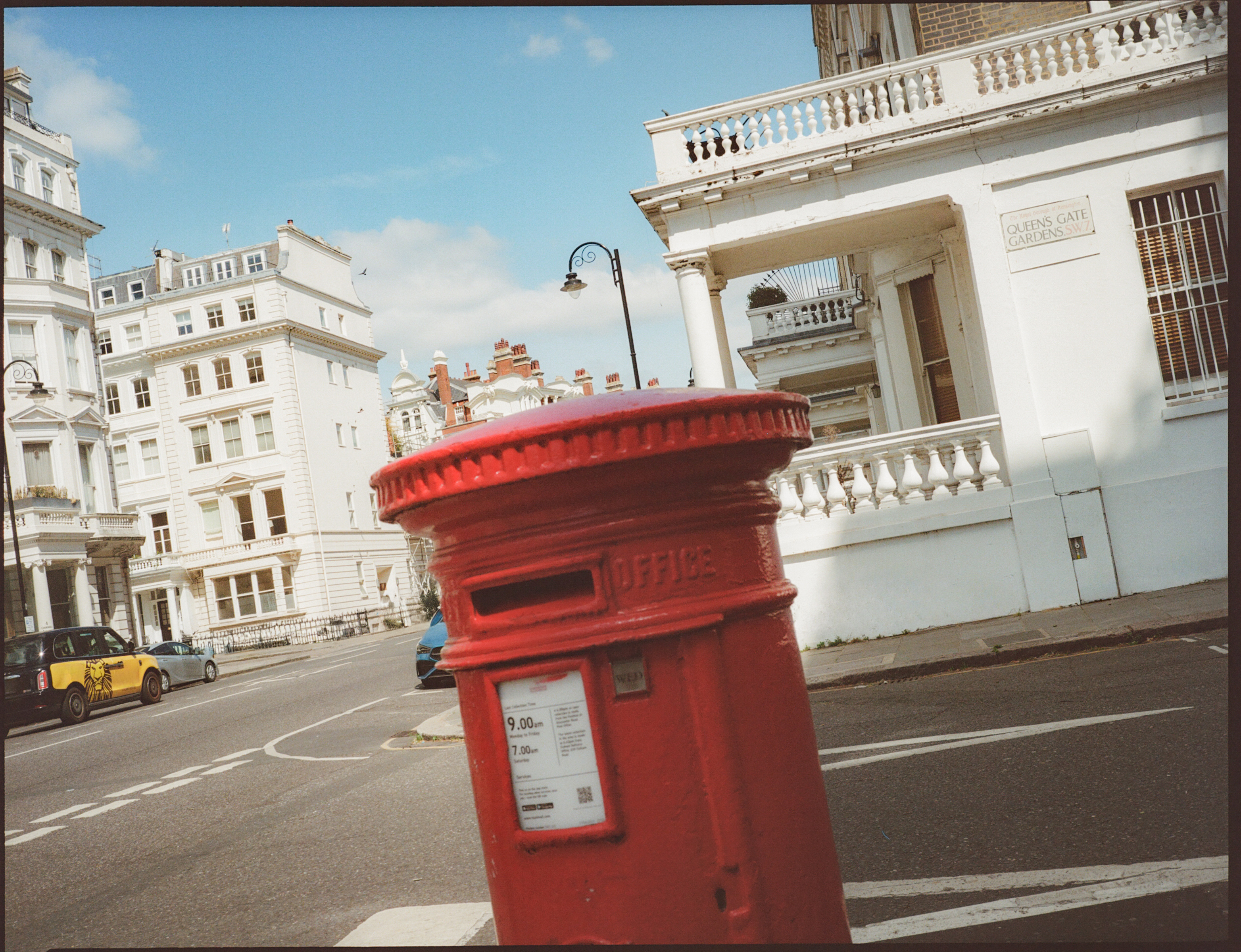 Buzón rojo de Londres en calle con edificios blancos, taxi y cielo azul.