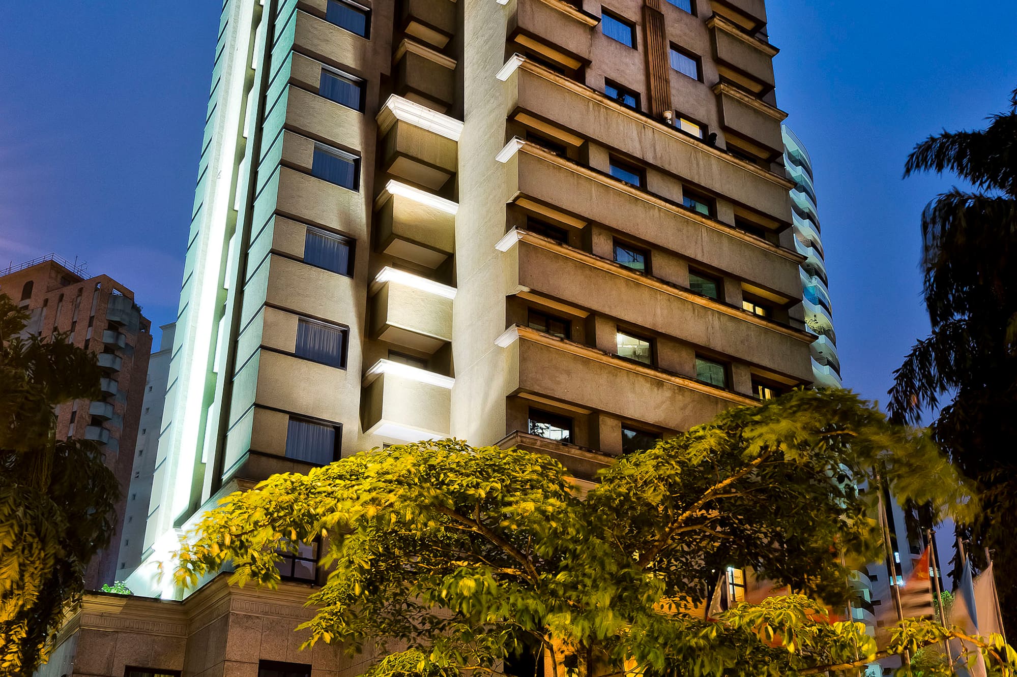 Modern high-rise hotel at night, illuminated tree, and blurred car light trails.