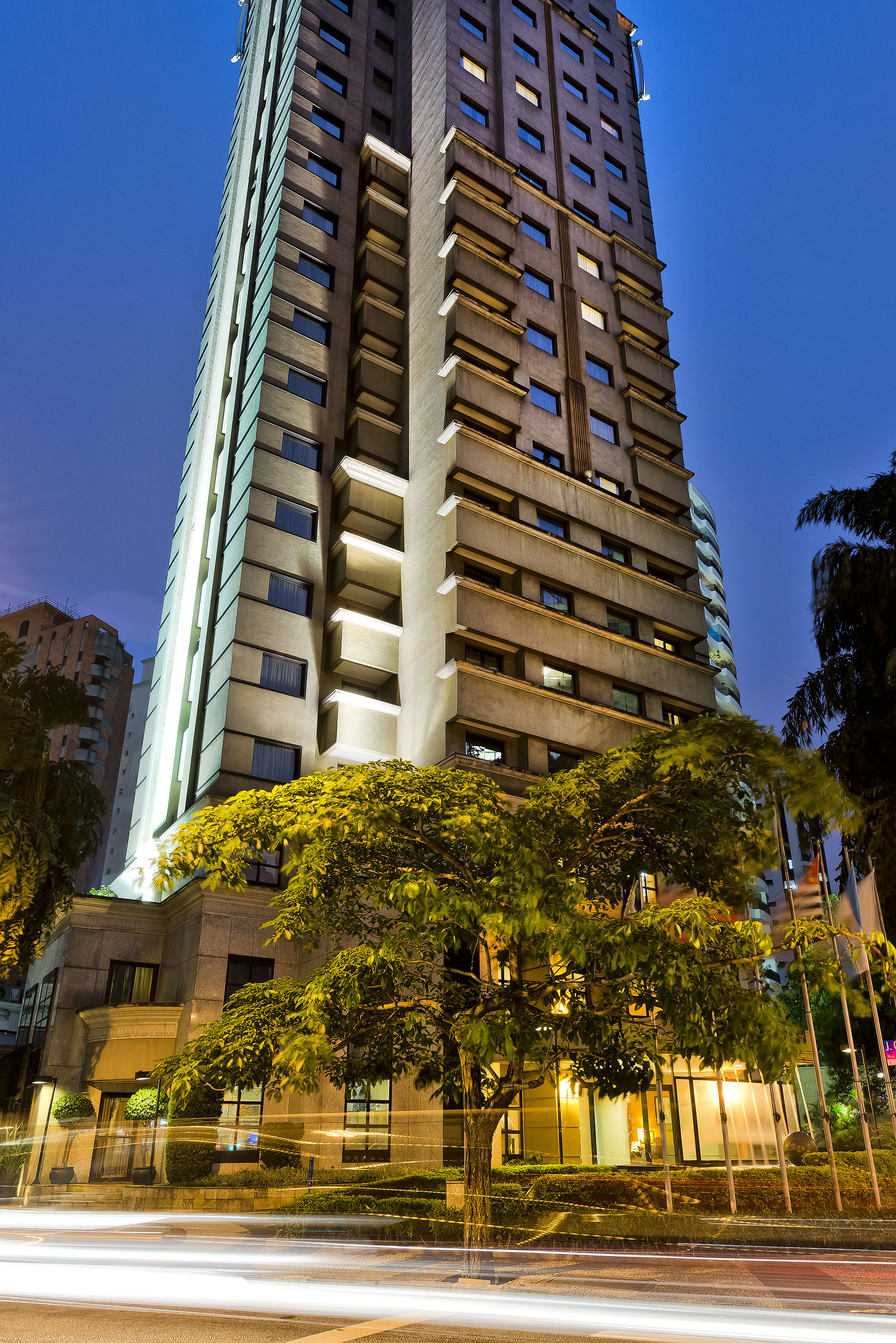 Modern high-rise hotel at night, illuminated tree, and blurred car light trails.