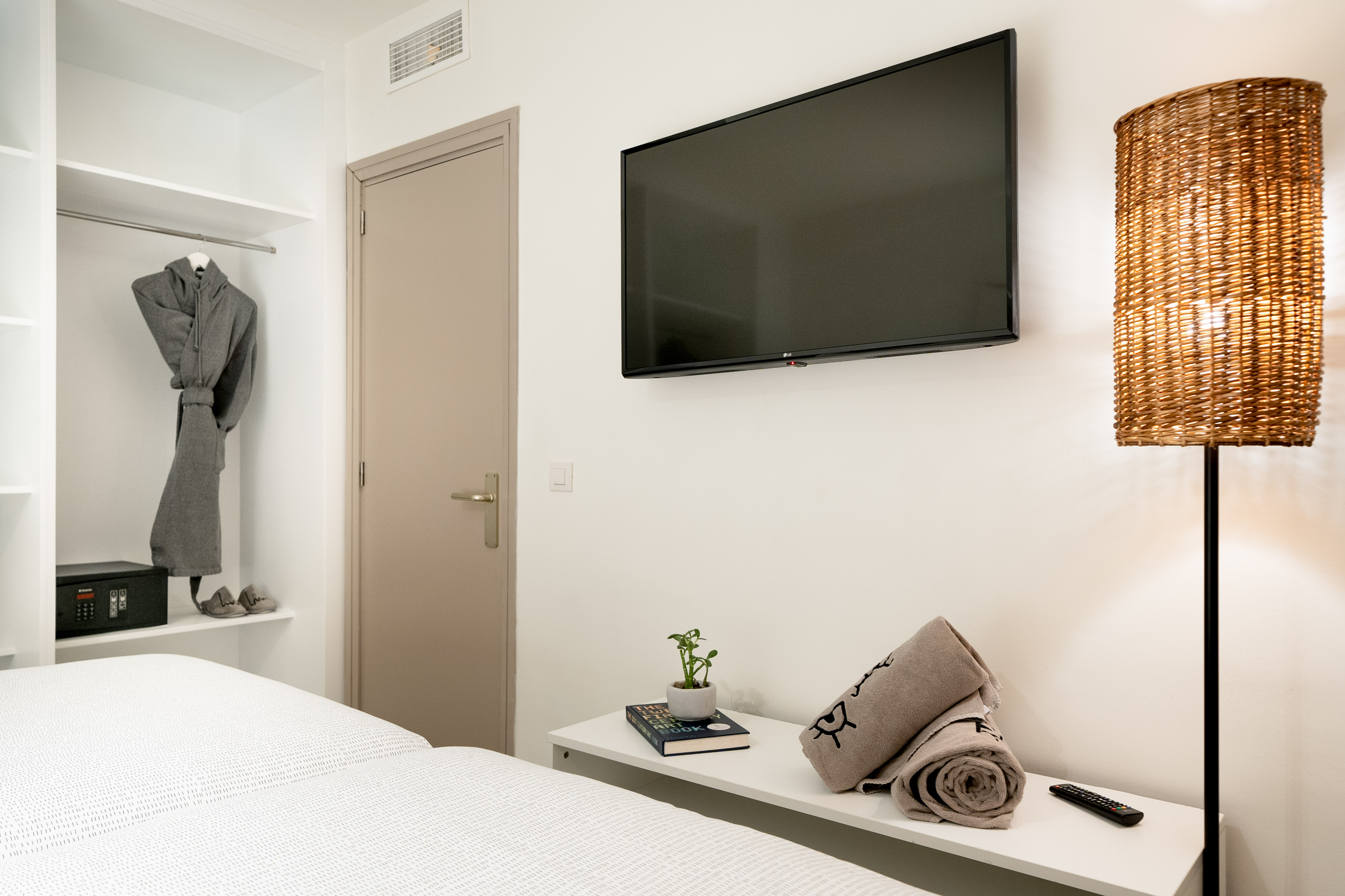 Modern hotel bedroom with wall-mounted TV, wicker lamp, open closet, and clean bed.