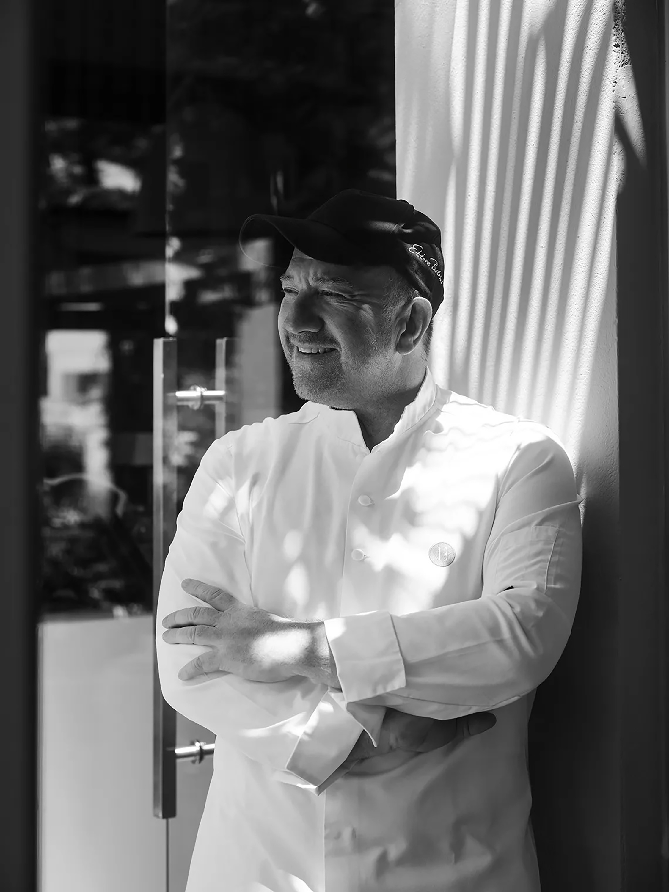 Smiling Chef Ettore Bal in B&W, wearing white jacket and cap, arms crossed.