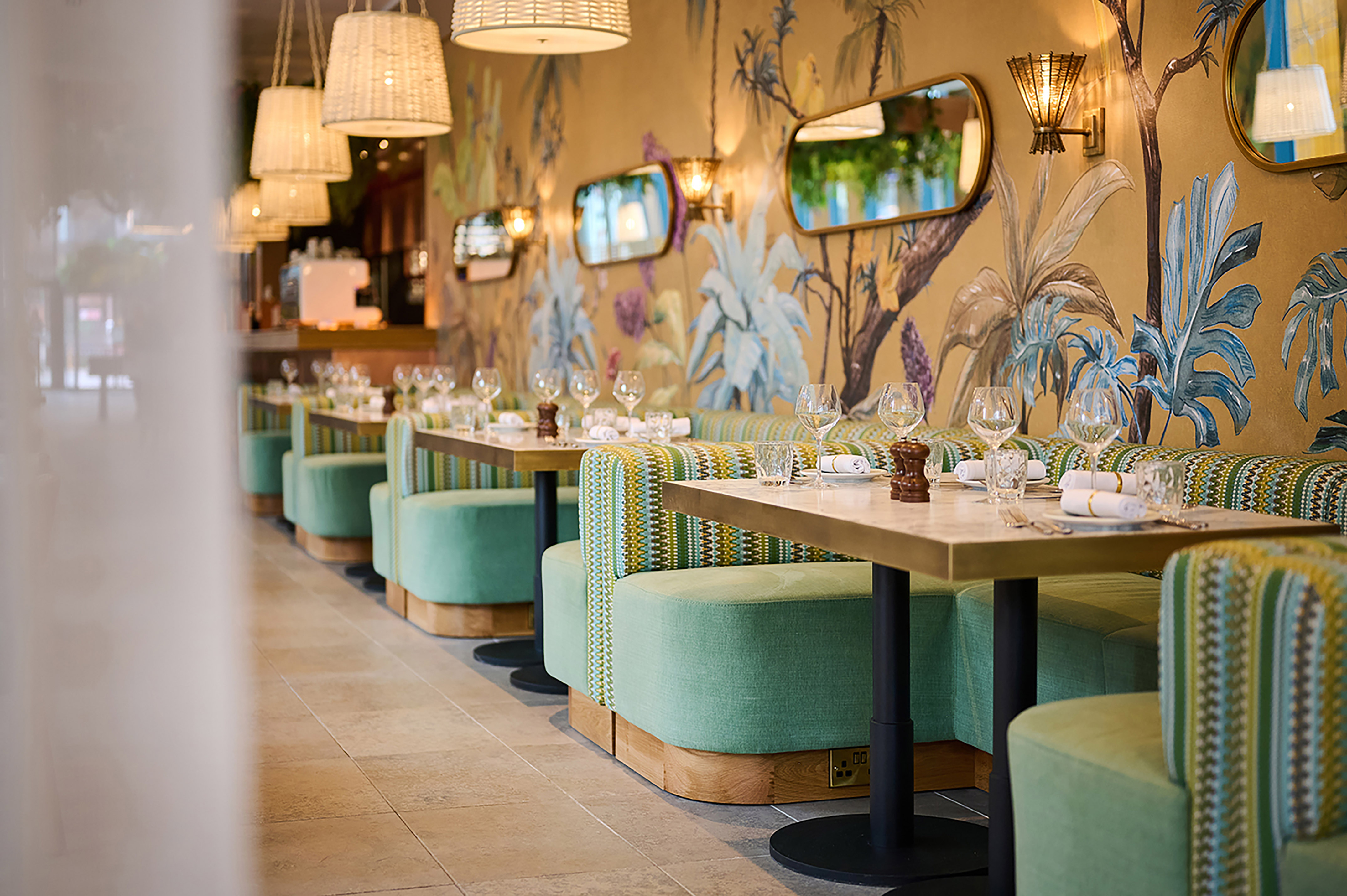 Restaurant dining room with green booths, tropical wallpaper, and set tables.