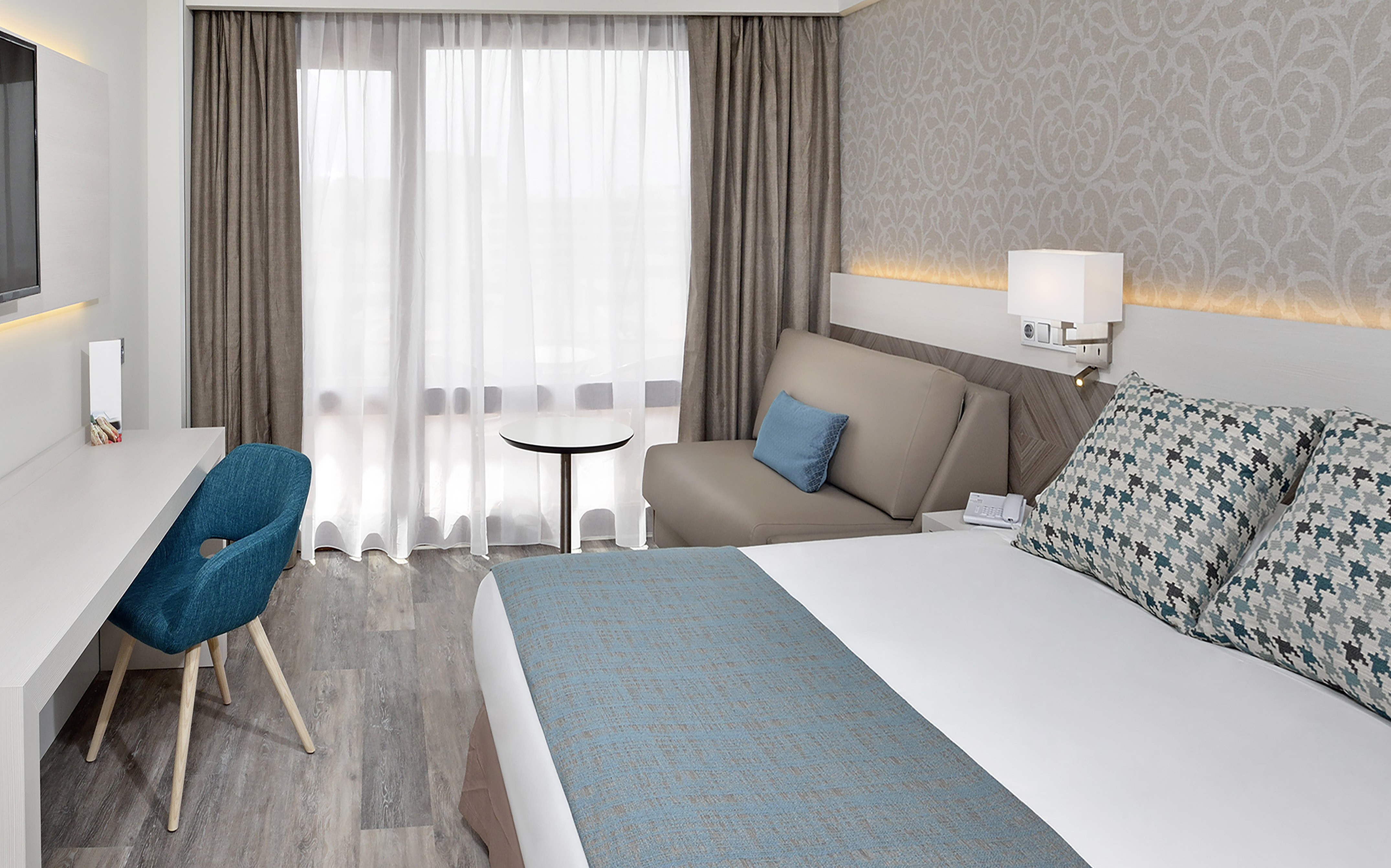 Modern hotel room with bed, desk, and seating area by a large window.