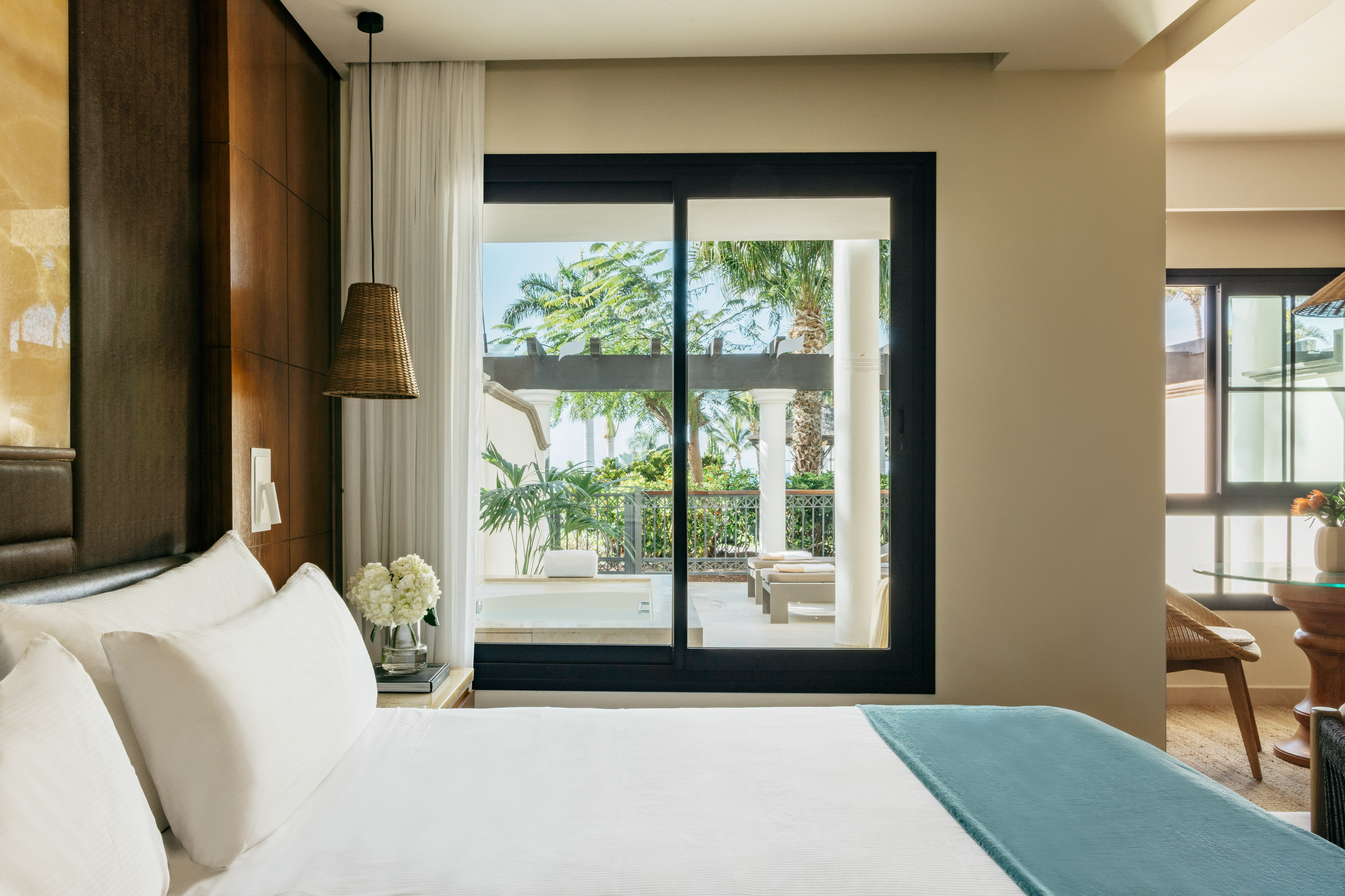 Resort bedroom with white bed, wicker lamp, and tropical garden view from balcony.