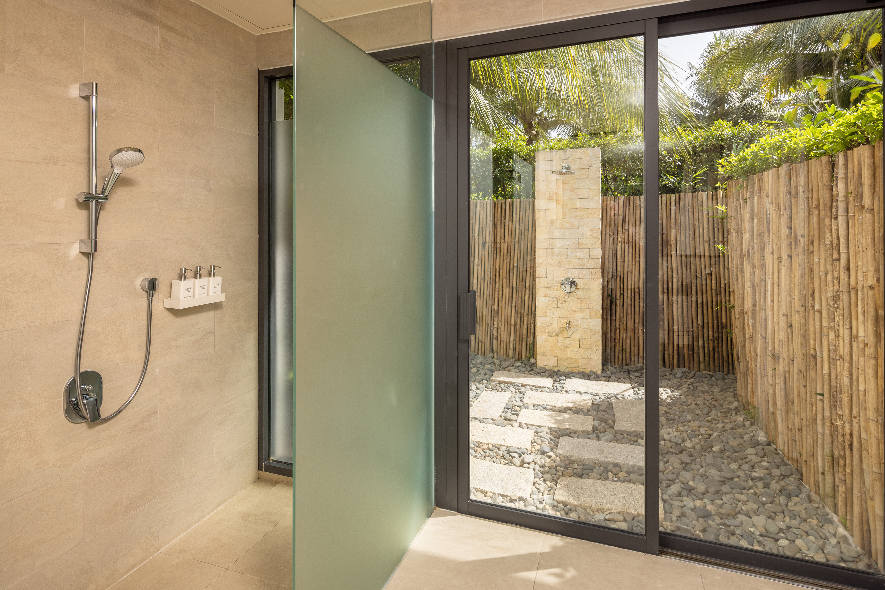 Indoor shower with glass door to tropical outdoor shower, bamboo, and pebbles.