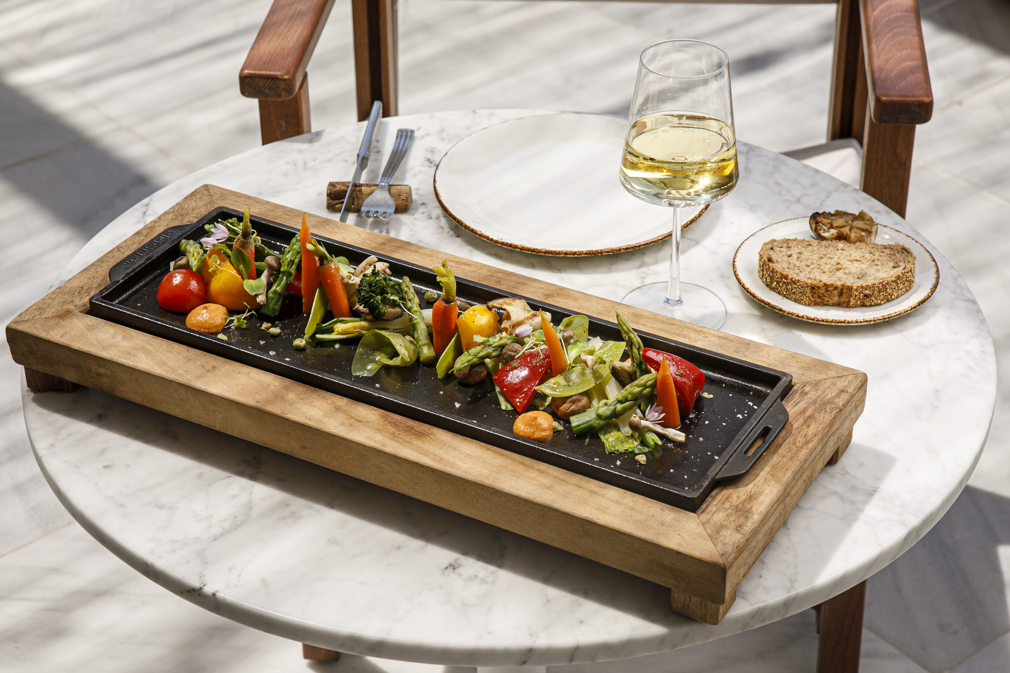 Colorful grilled vegetables, white wine, and whole grain bread on marble table.