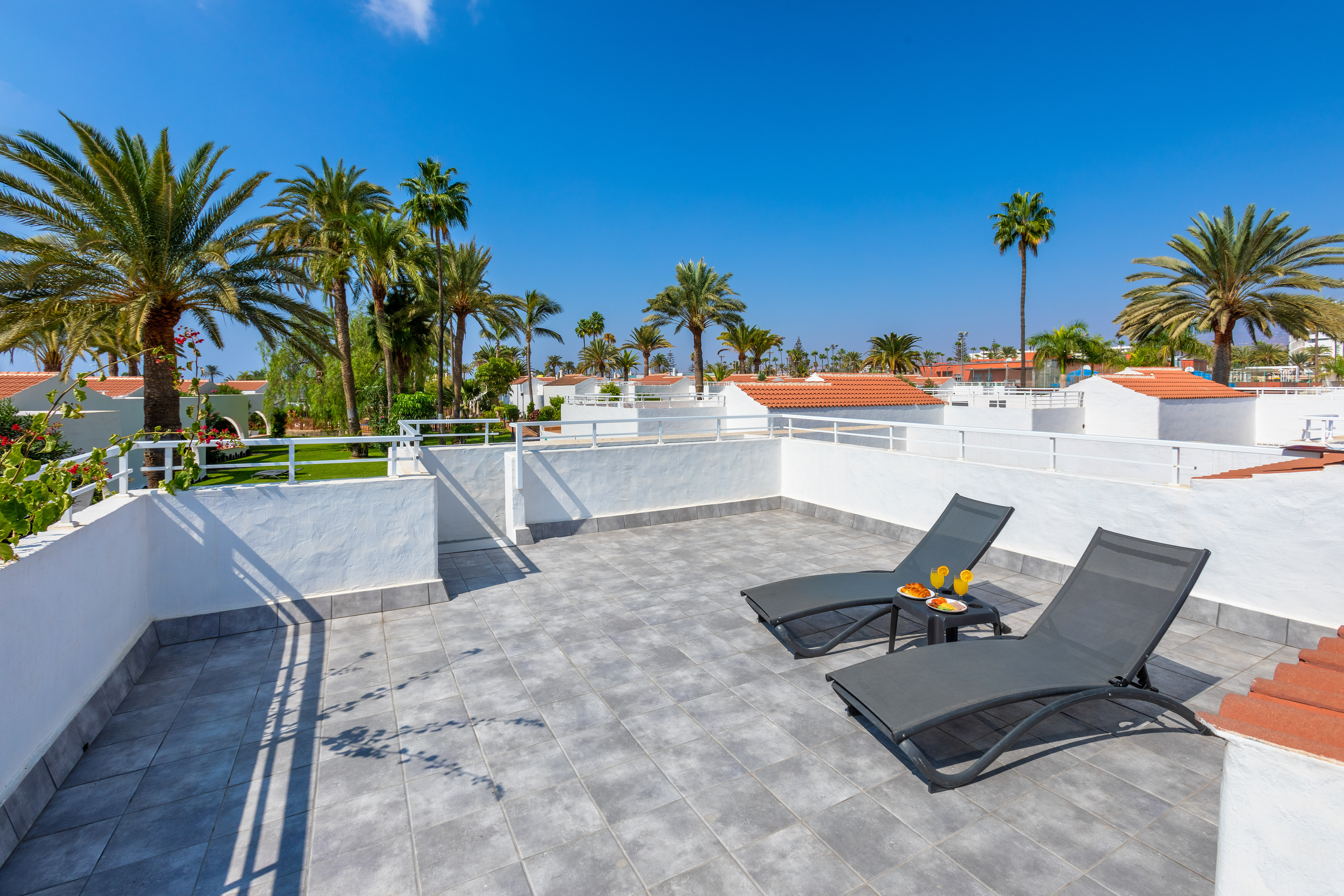 Relaxing resort rooftop terrace with sun loungers, juice, and palm trees.