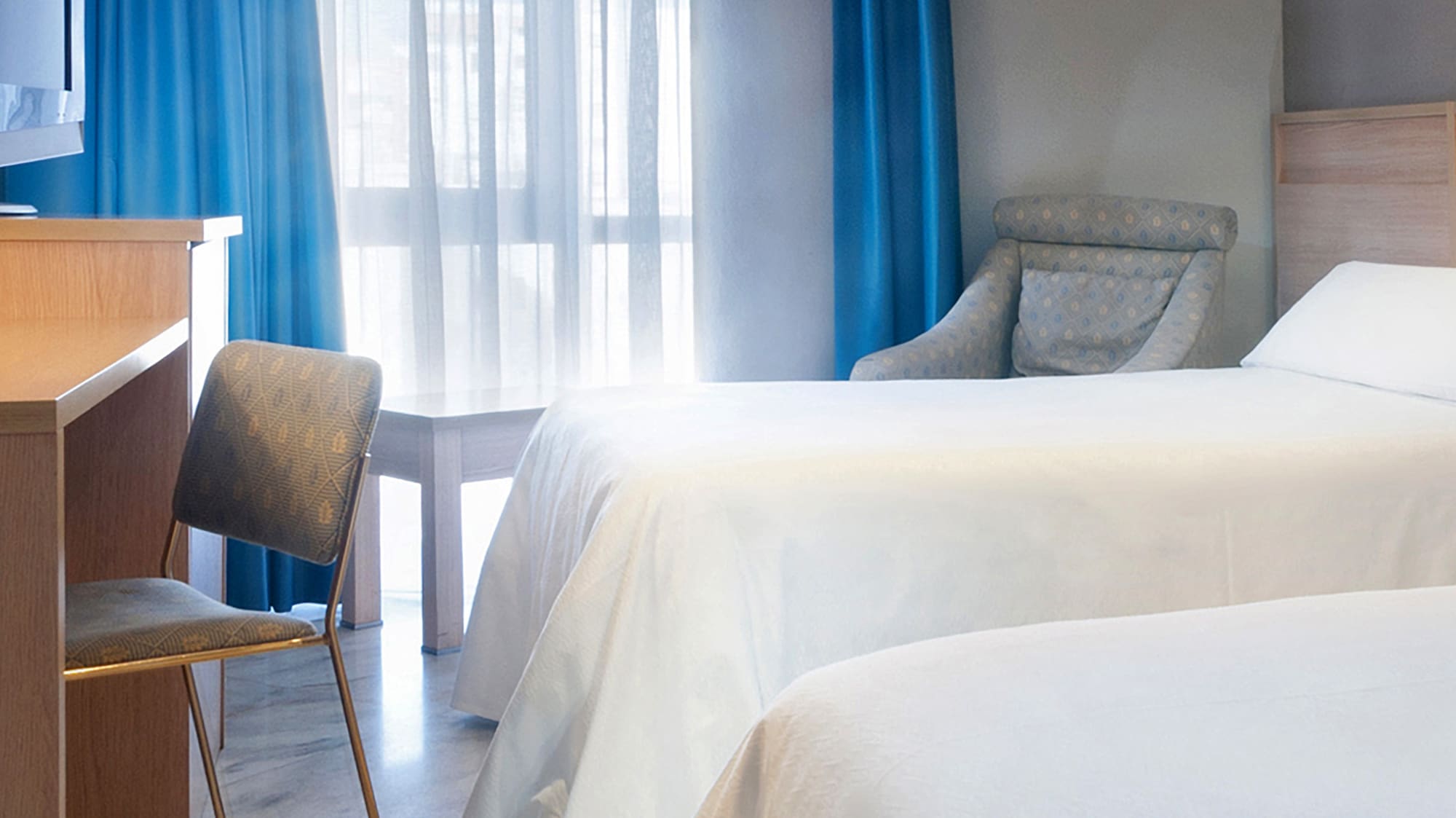 Hotel guest room with twin beds, blue curtains, desk, TV, and marble floor.