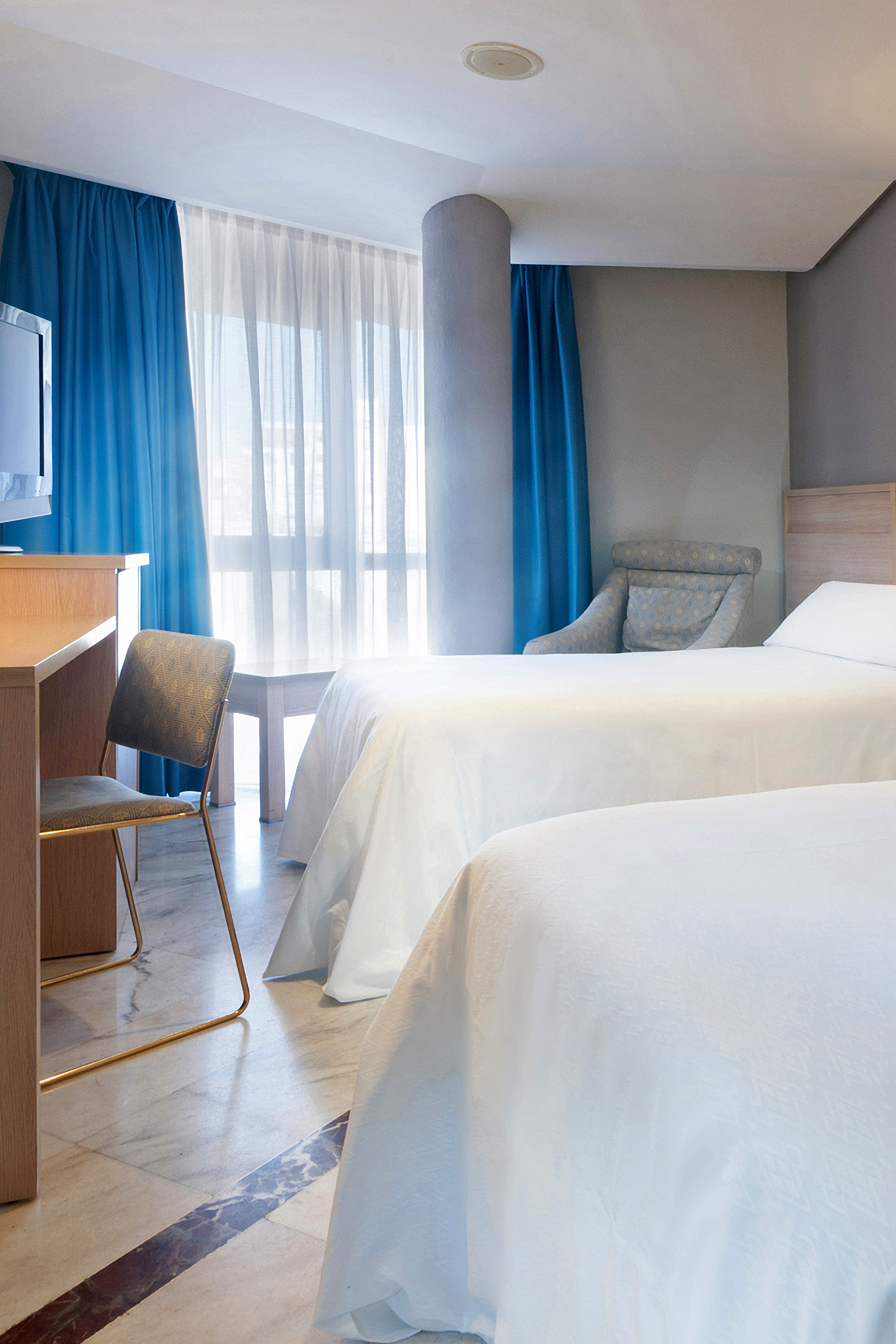 Hotel guest room with twin beds, blue curtains, desk, TV, and marble floor.