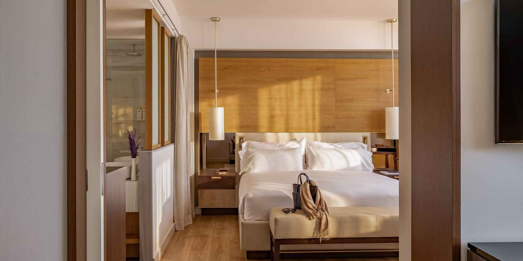 Sunlit modern hotel bedroom with king bed, wooden decor, and ensuite bath.