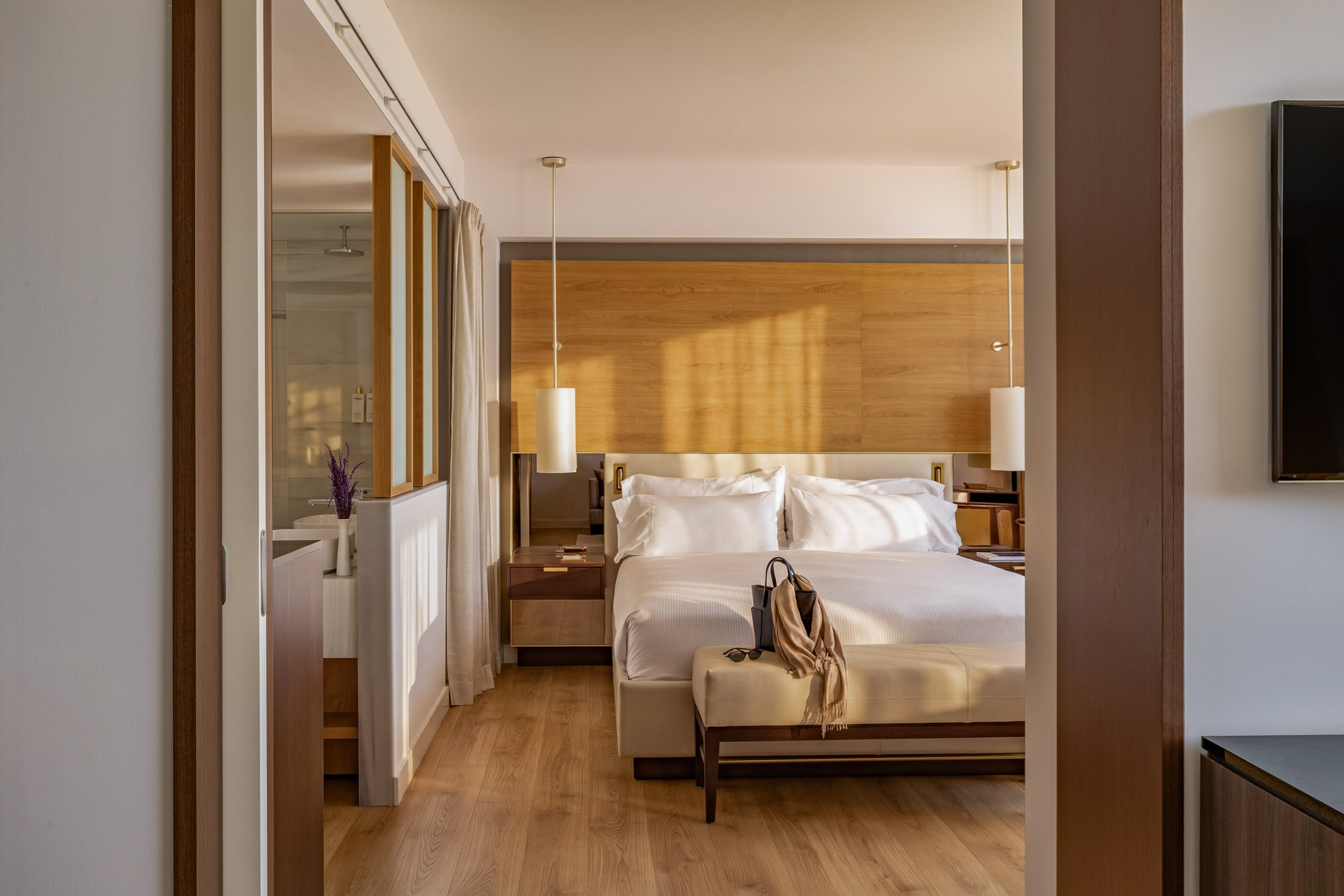 Sunlit modern hotel bedroom with king bed, wooden decor, and ensuite bath.