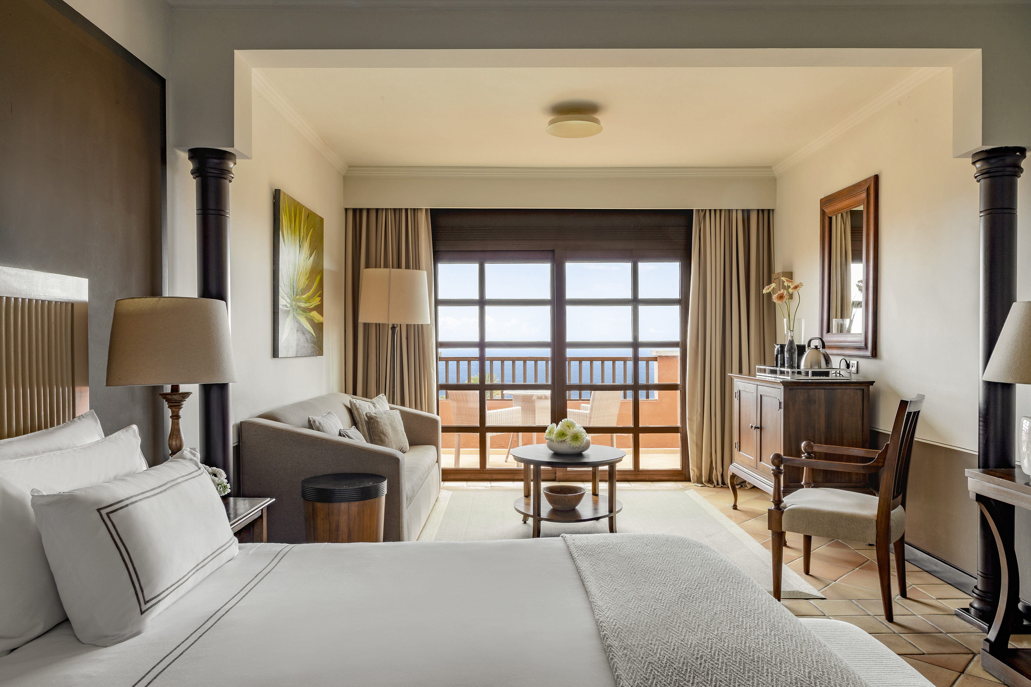 Hotel suite with large bed, sitting area, and ocean view from private balcony.