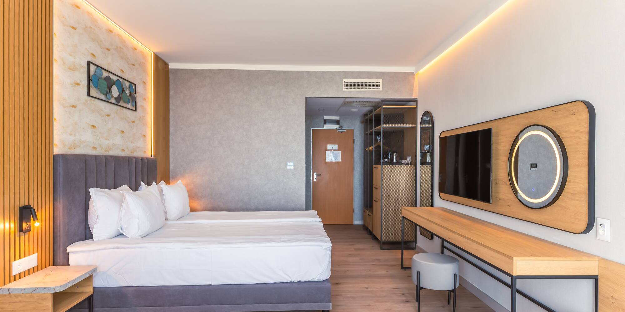 Modern hotel room with bed, wood floor, ambient lighting, TV, and vanity.