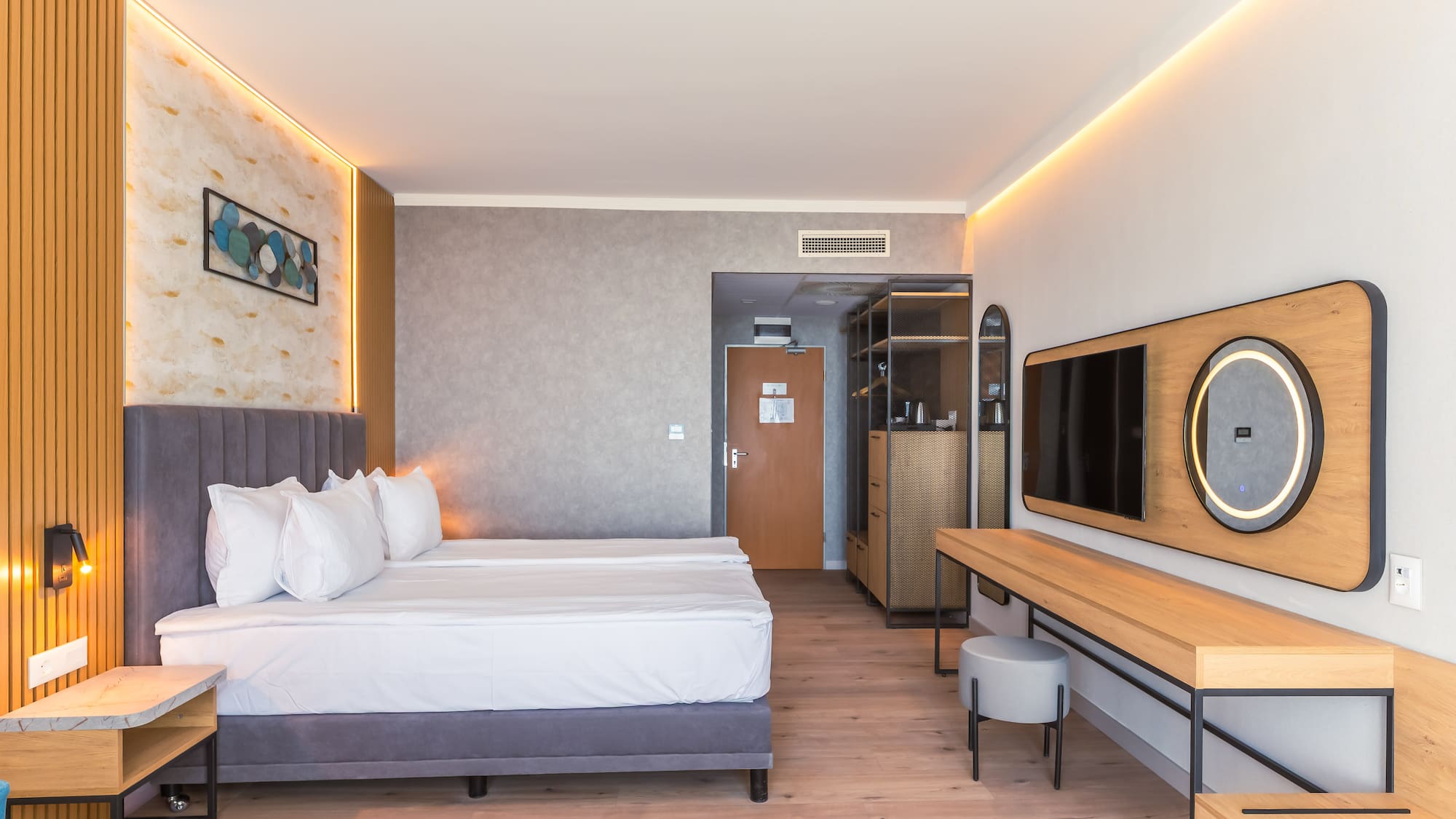 Modern hotel room with bed, wood floor, ambient lighting, TV, and vanity.