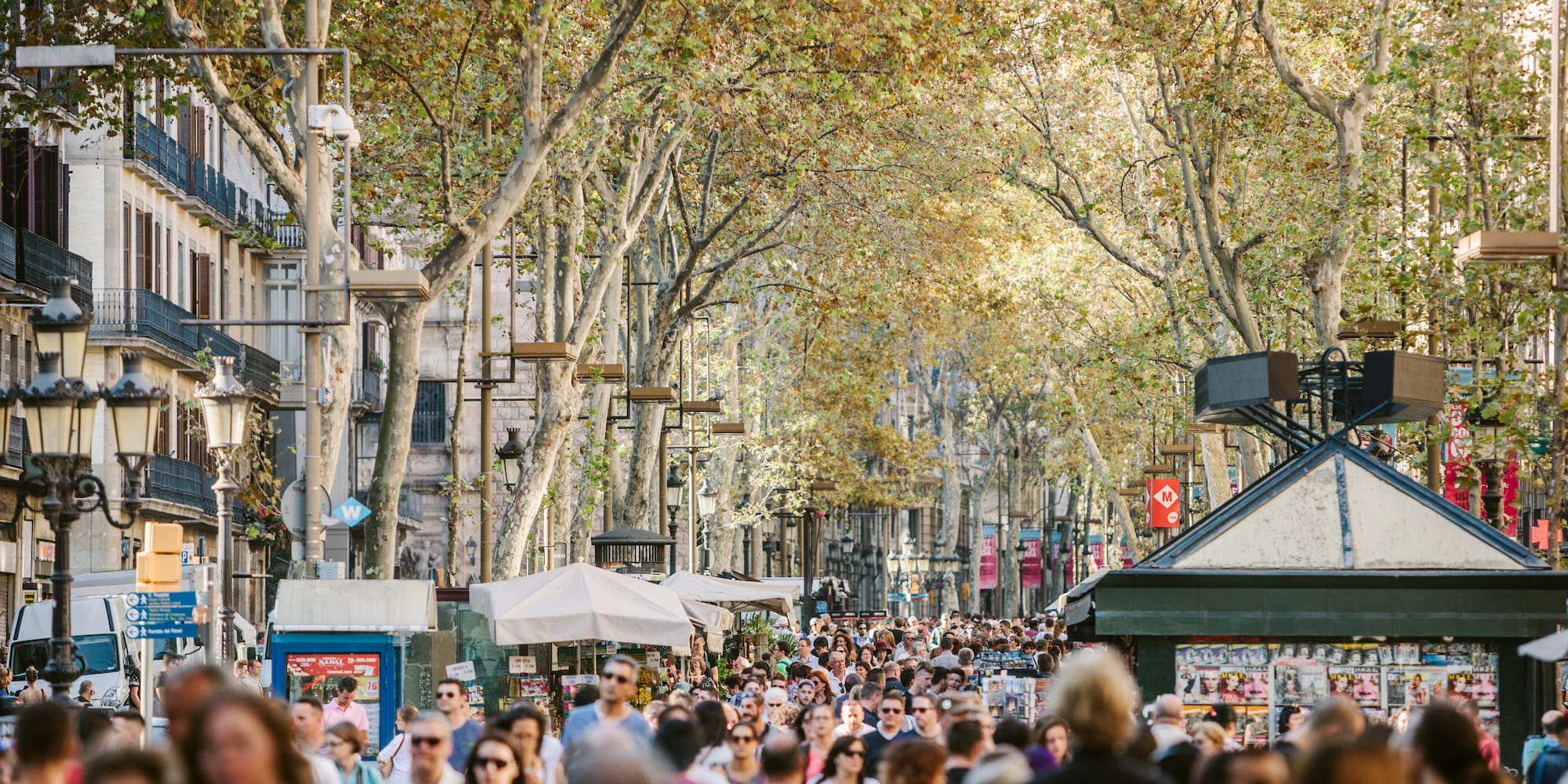 Paseo concurrido en La Rambla, Barcelona, con árboles otoñales y quioscos.