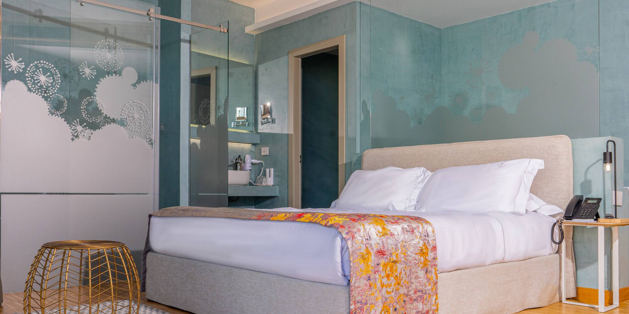 Modern hotel bedroom with large bed, teal walls, and frosted glass shower.