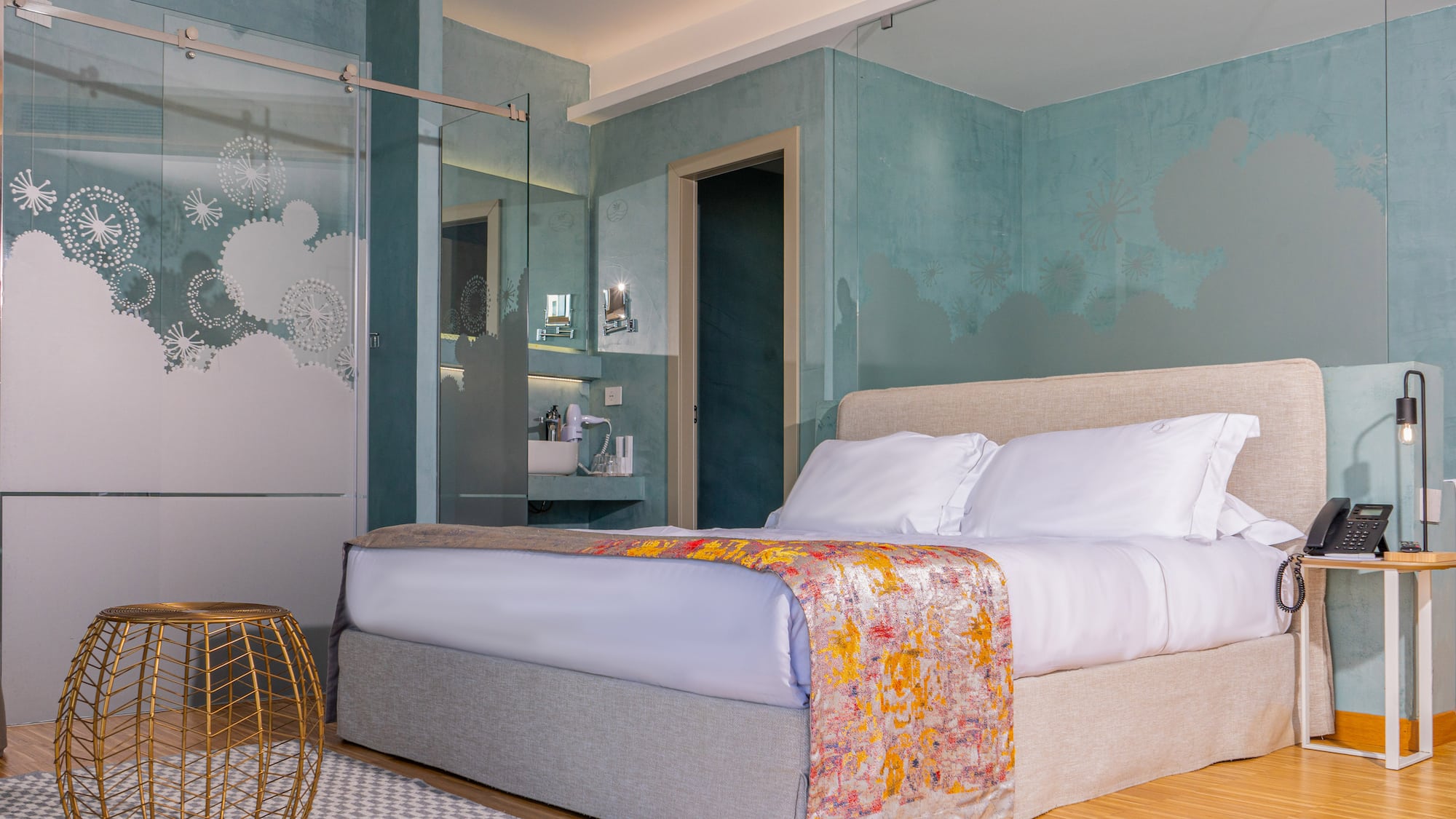 Modern hotel bedroom with large bed, teal walls, and frosted glass shower.