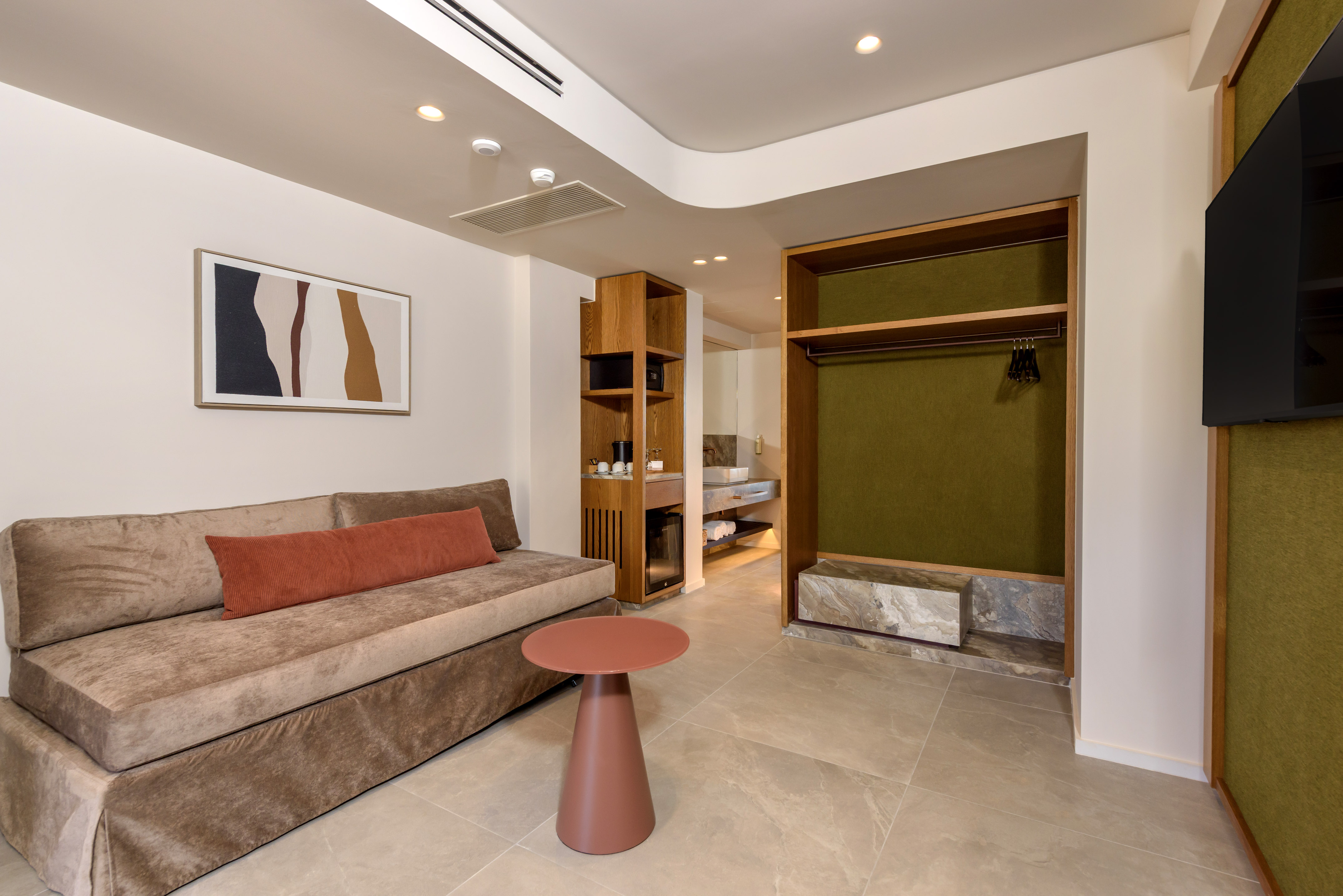 Modern hotel room with sofa, side table, kitchenette, and open wardrobe.