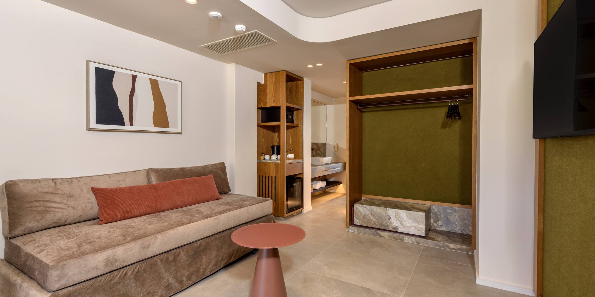 Modern hotel room with sofa, side table, kitchenette, and open wardrobe.
