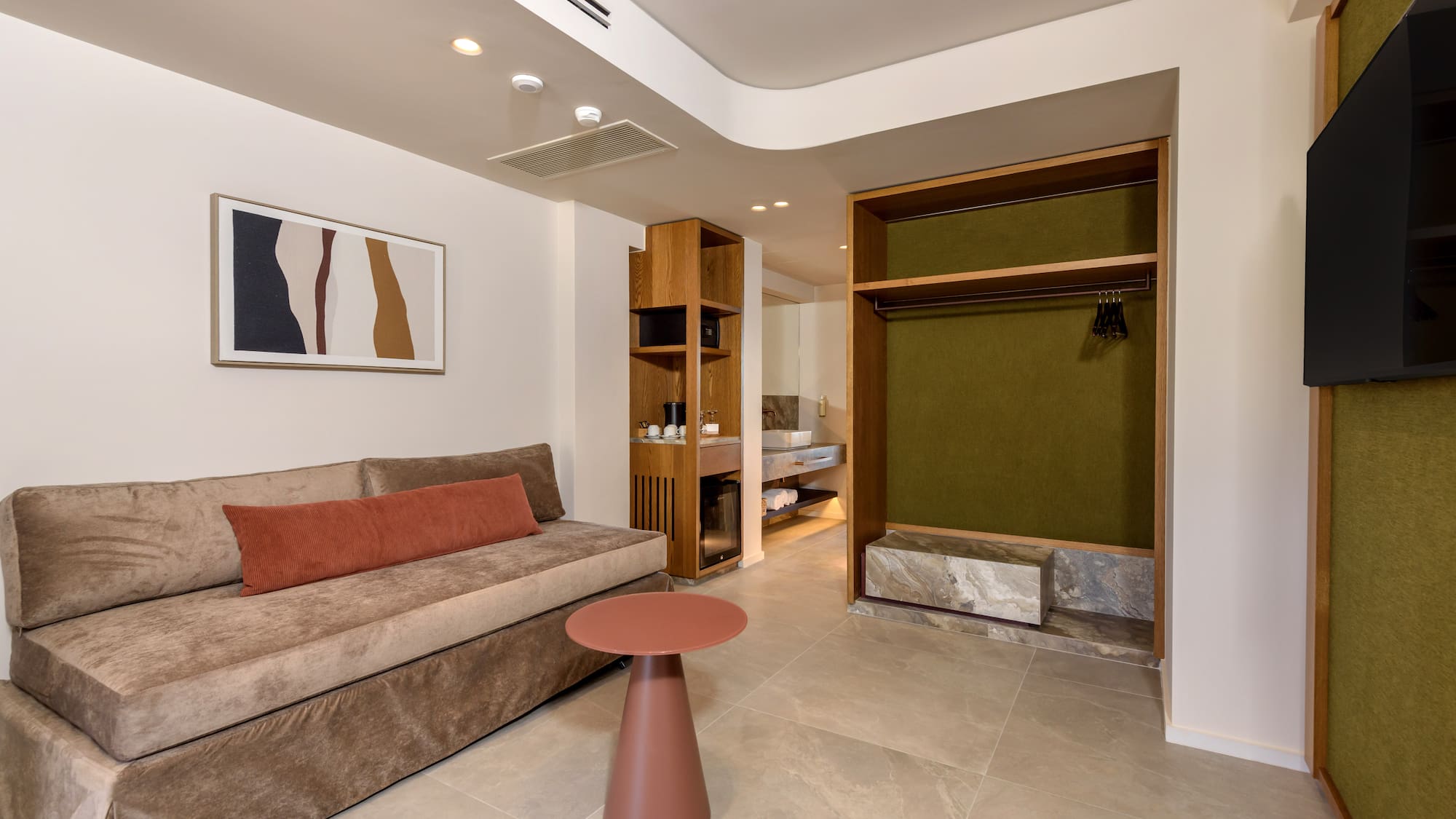 Modern hotel room with sofa, side table, kitchenette, and open wardrobe.